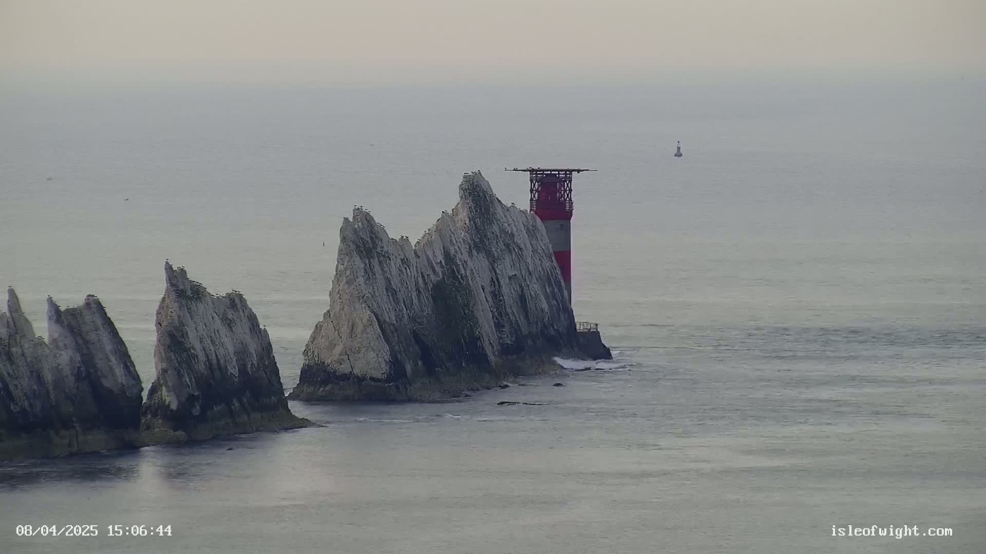 A red and white lighthouse stands atop a cluster of tall, white, rocky cliffs surrounded by a calm, gray sea under an overcast sky.