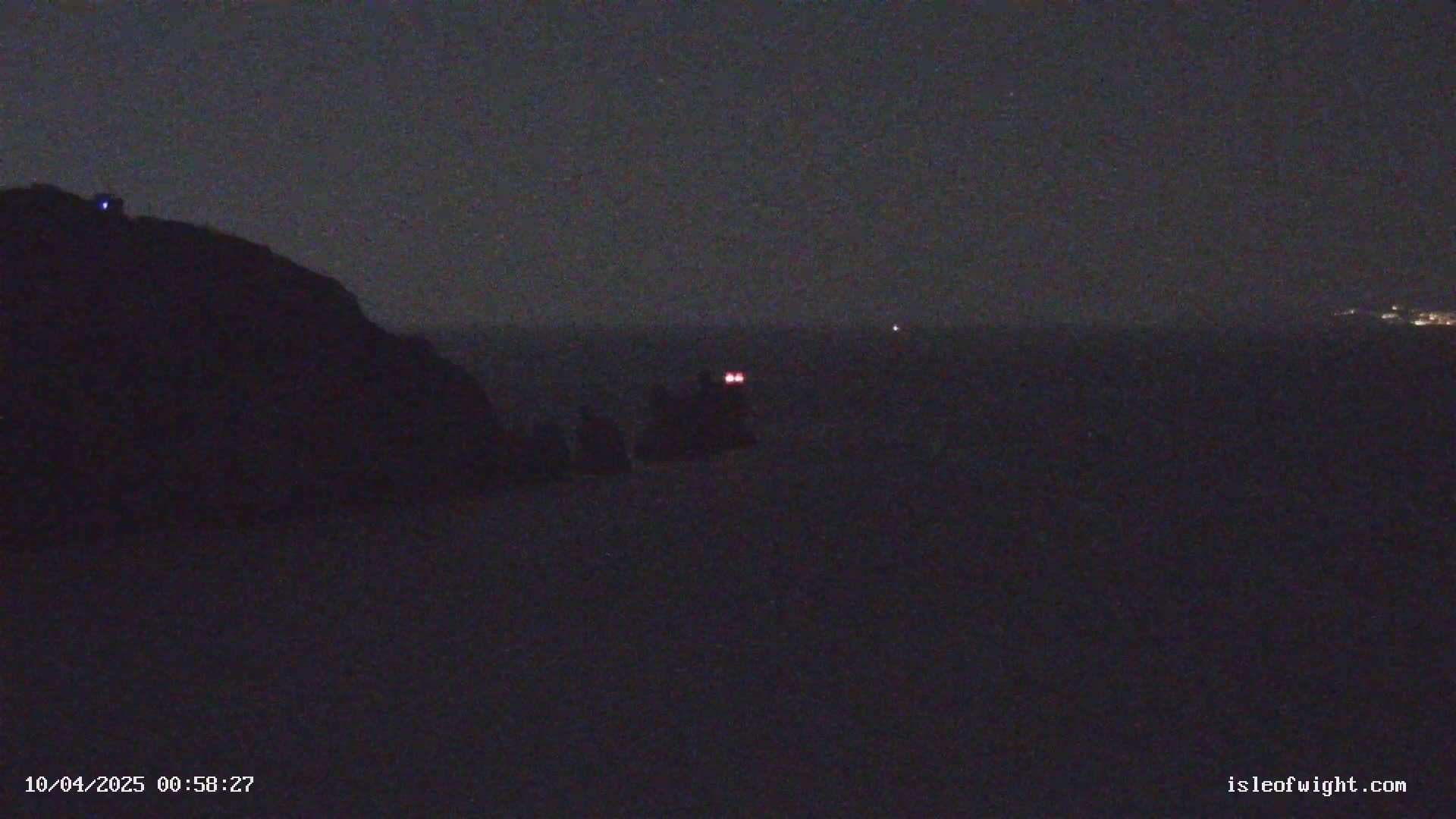A dark, nighttime scene shows a large, dark landmass on the left, a body of water in the center with a small vessel showing two red lights, and a faint light source on the horizon to the right.