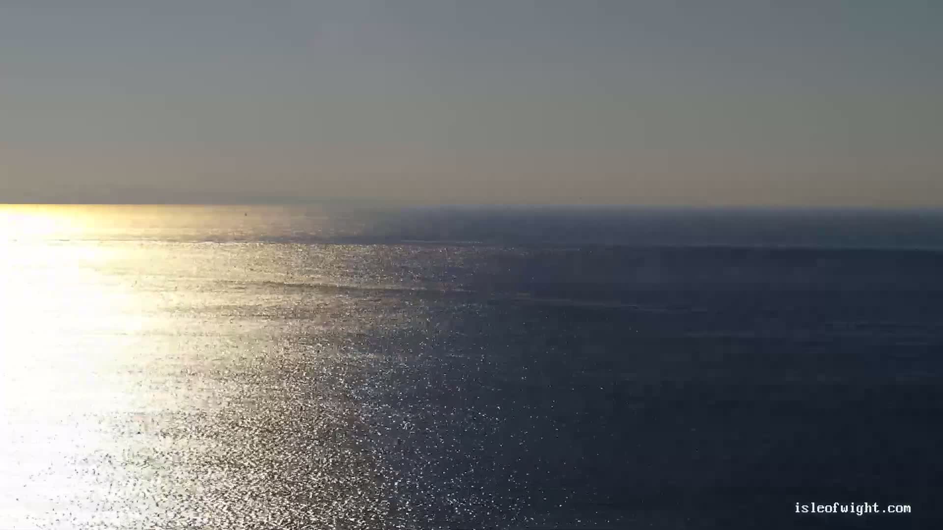 The sun shines brightly on a calm, expansive ocean, its surface shimmering with a multitude of glittering reflections.