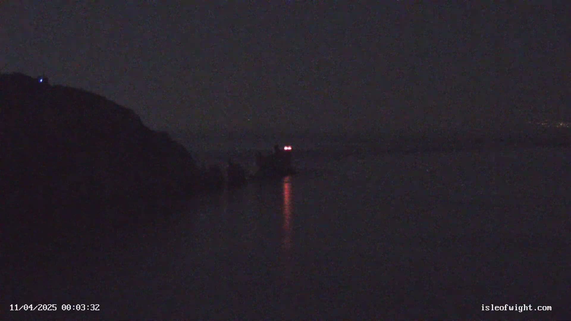 A dark, nighttime scene shows a body of water with a structure exhibiting two red lights in the distance, a dark landmass to the left, and a hazy or foggy atmosphere.