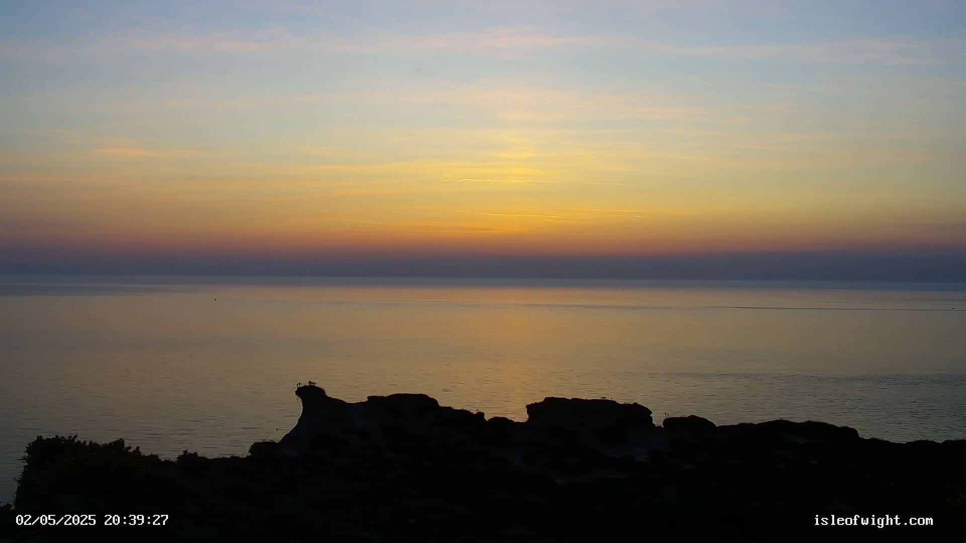 A sunset over a calm sea, viewed from a rocky cliff silhouetted against the colorful sky.