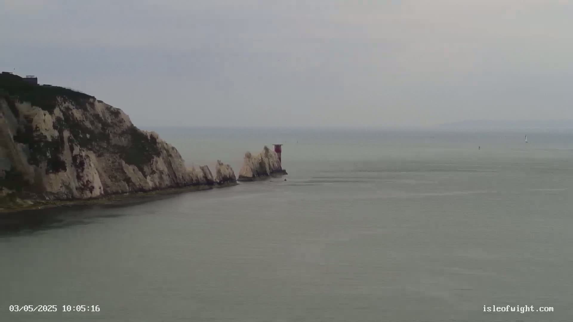White cliffs meet a calm, gray sea, featuring a small, red lighthouse on a rocky outcrop.