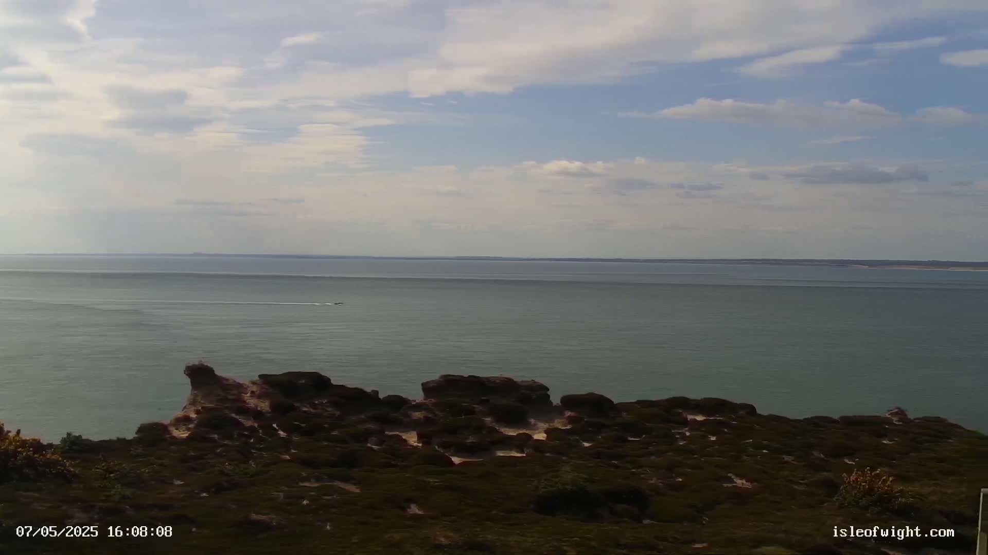 A partly cloudy sky overlooks a calm sea with a small vessel in the distance, and dark-green shrubbery covers a rocky, coastal outcropping in the foreground.