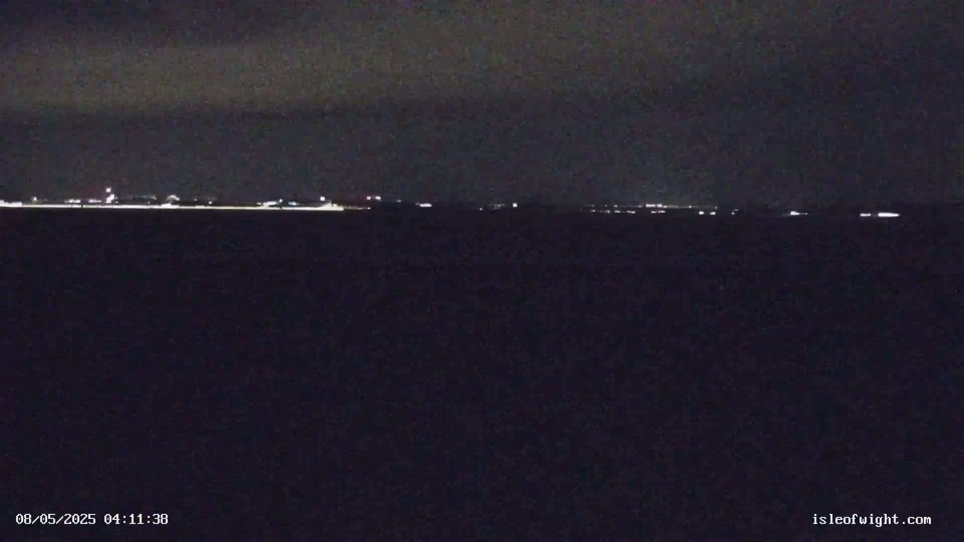 A dark, nighttime scene shows a distant line of faint lights across a dark body of water under a cloudy sky.