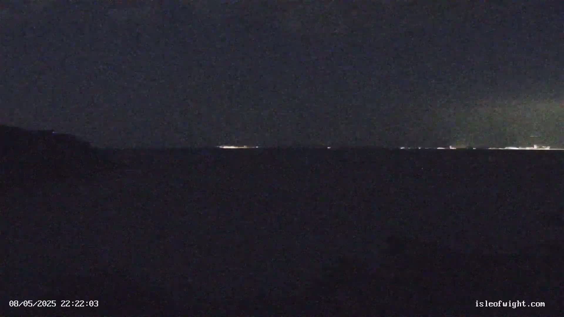 A dark, nighttime view of a body of water shows a faint line of lights on the horizon under a mostly clear, dark sky.
