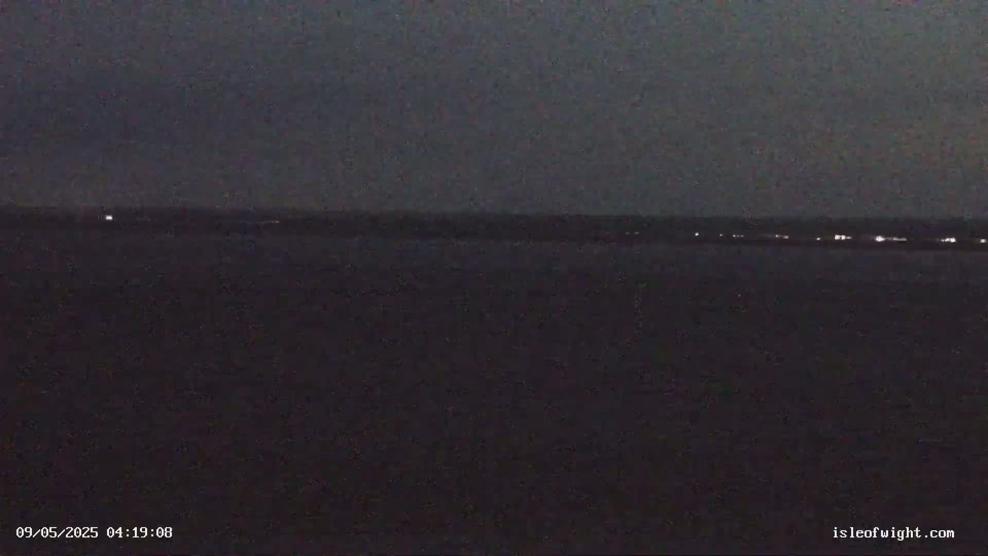 A dark, nighttime scene shows a body of water with a distant shoreline illuminated by faint lights under a mostly clear, dark sky.