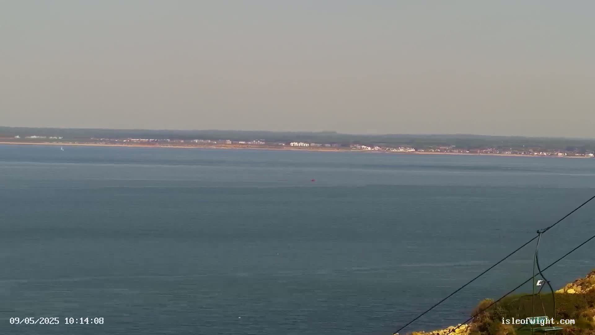 A calm, hazy ocean view shows a distant shoreline with buildings, a small boat on the water, and a section of cable lift in the foreground.