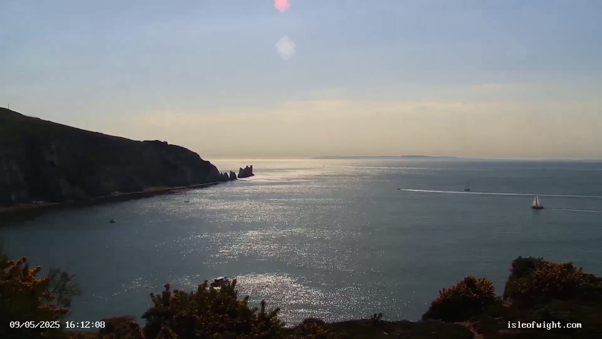A sunny, calm ocean scene shows a rocky cliffside on the left, two sailboats in the distance, and sparkling water reflecting sunlight.