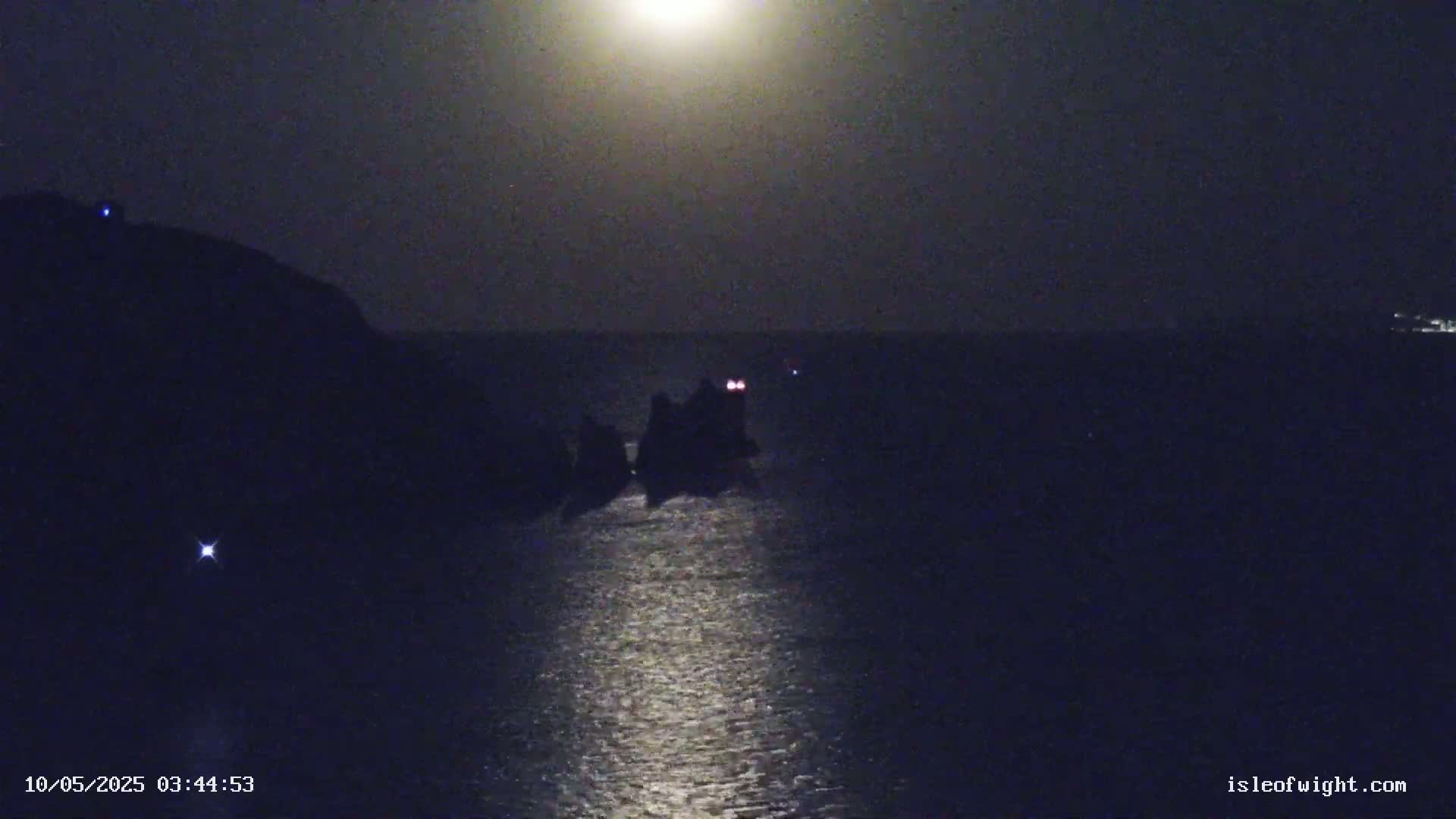 Under a bright moonlit night sky, dark, rocky landmasses jut out into a calm sea, reflecting the moon's light.