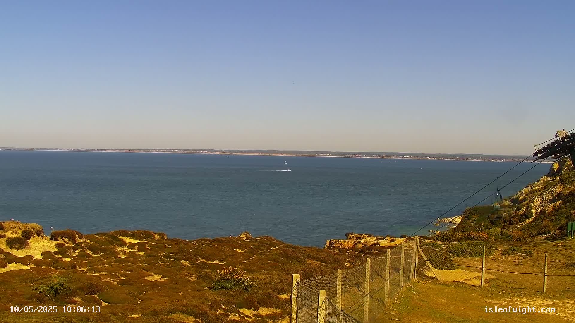 A calm, sunny day overlooks a vast expanse of ocean from a clifftop, featuring a small boat in the distance and a chain-link fence in the foreground.