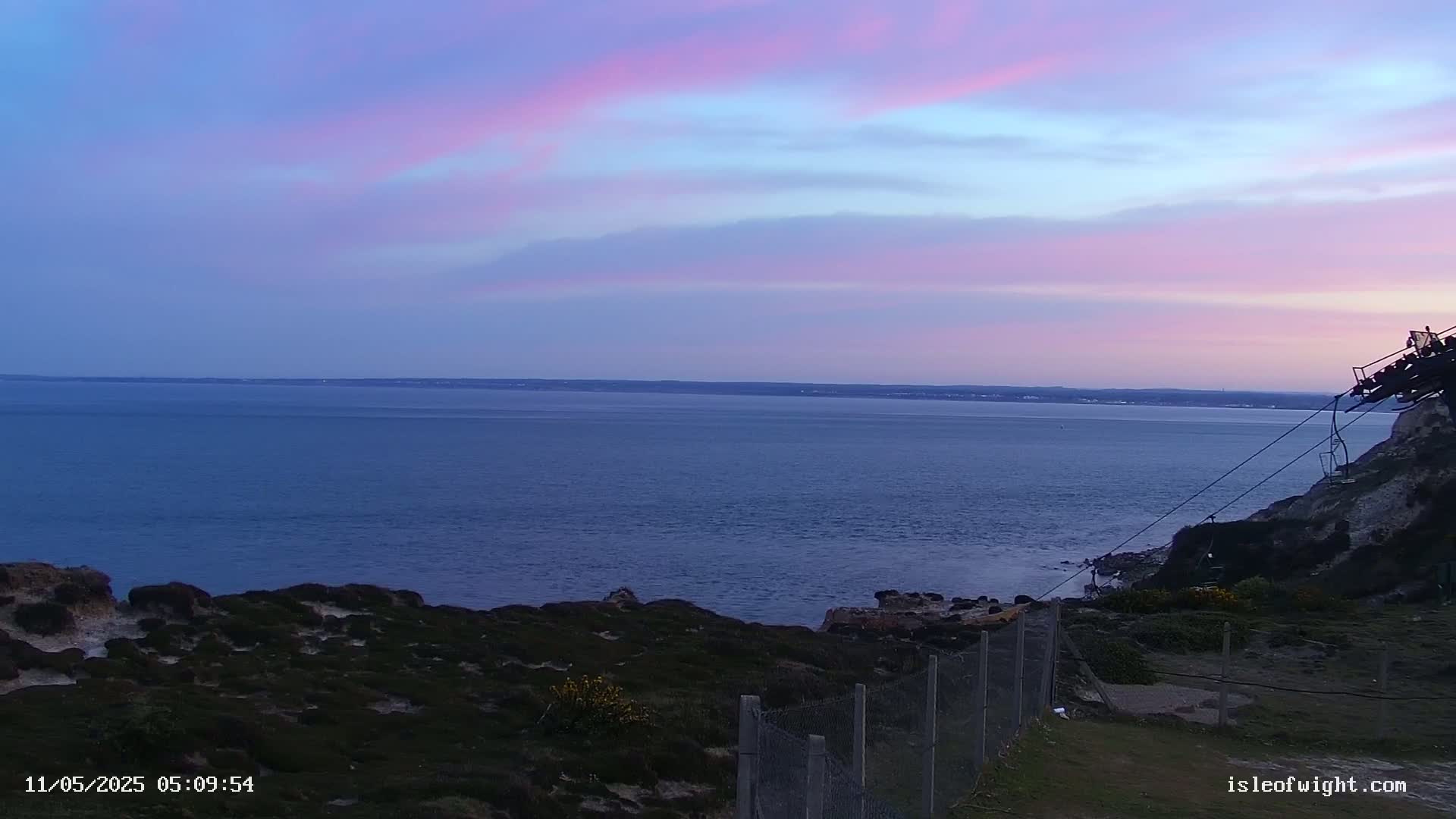 A calm sea stretches to a distant land under a pastel-colored sunset sky, viewed from a cliff top with sparse vegetation and a wire fence.