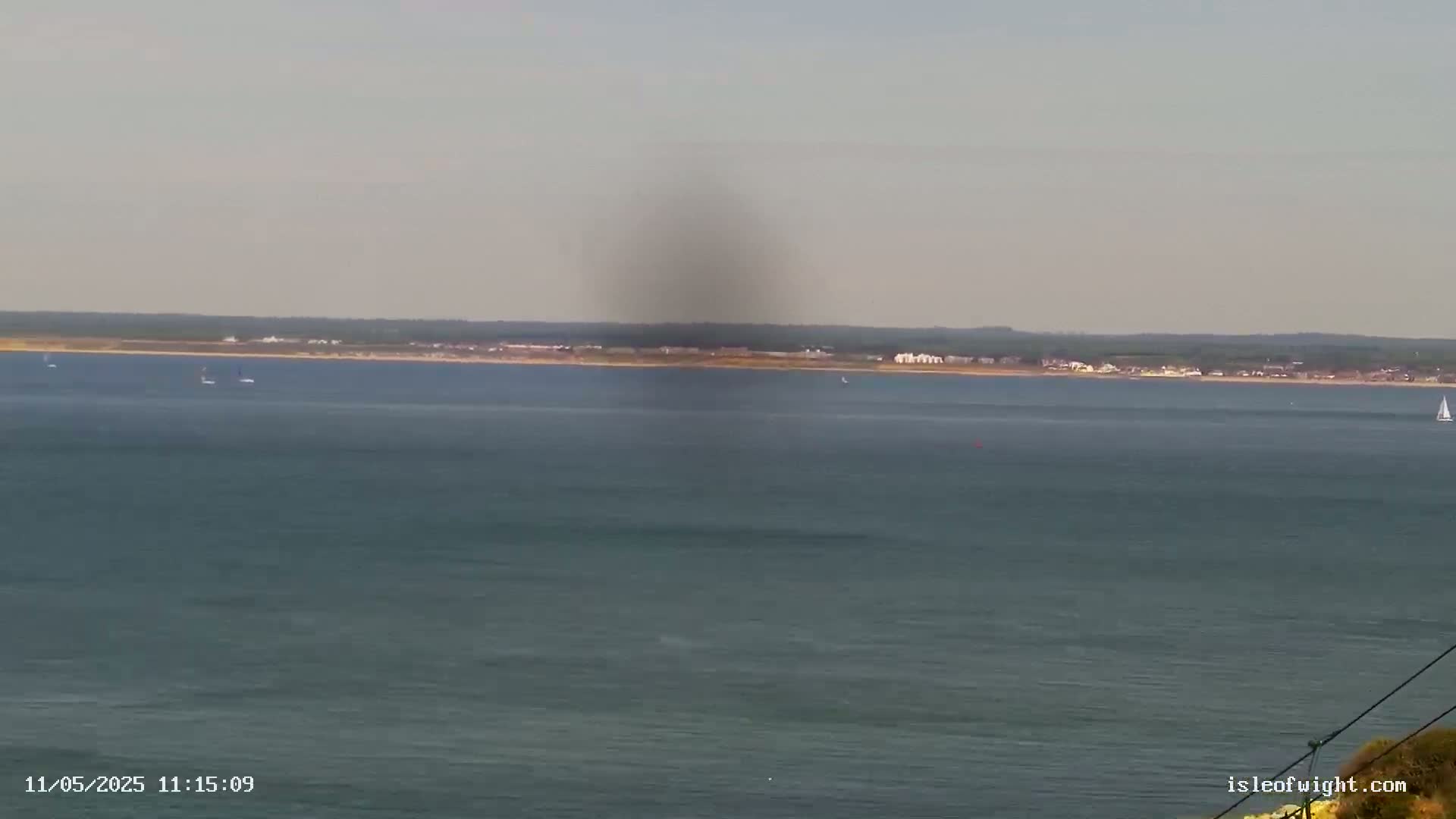 A hazy, calm ocean scene shows a distant shoreline with a few sailboats and a large dark smudge in the sky.