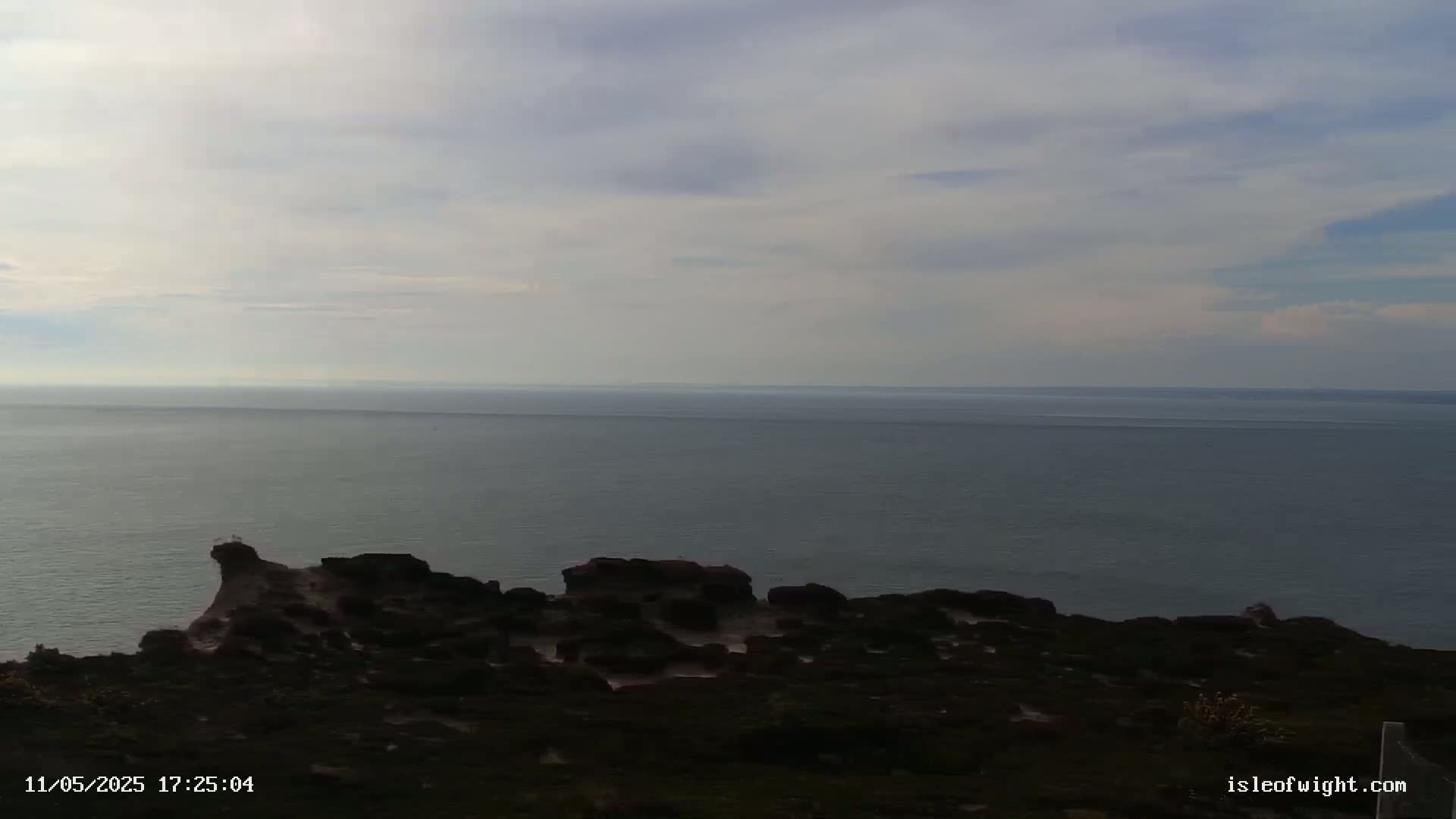 A rocky, dark coastline overlooks a calm, grey sea under a mostly cloudy sky.