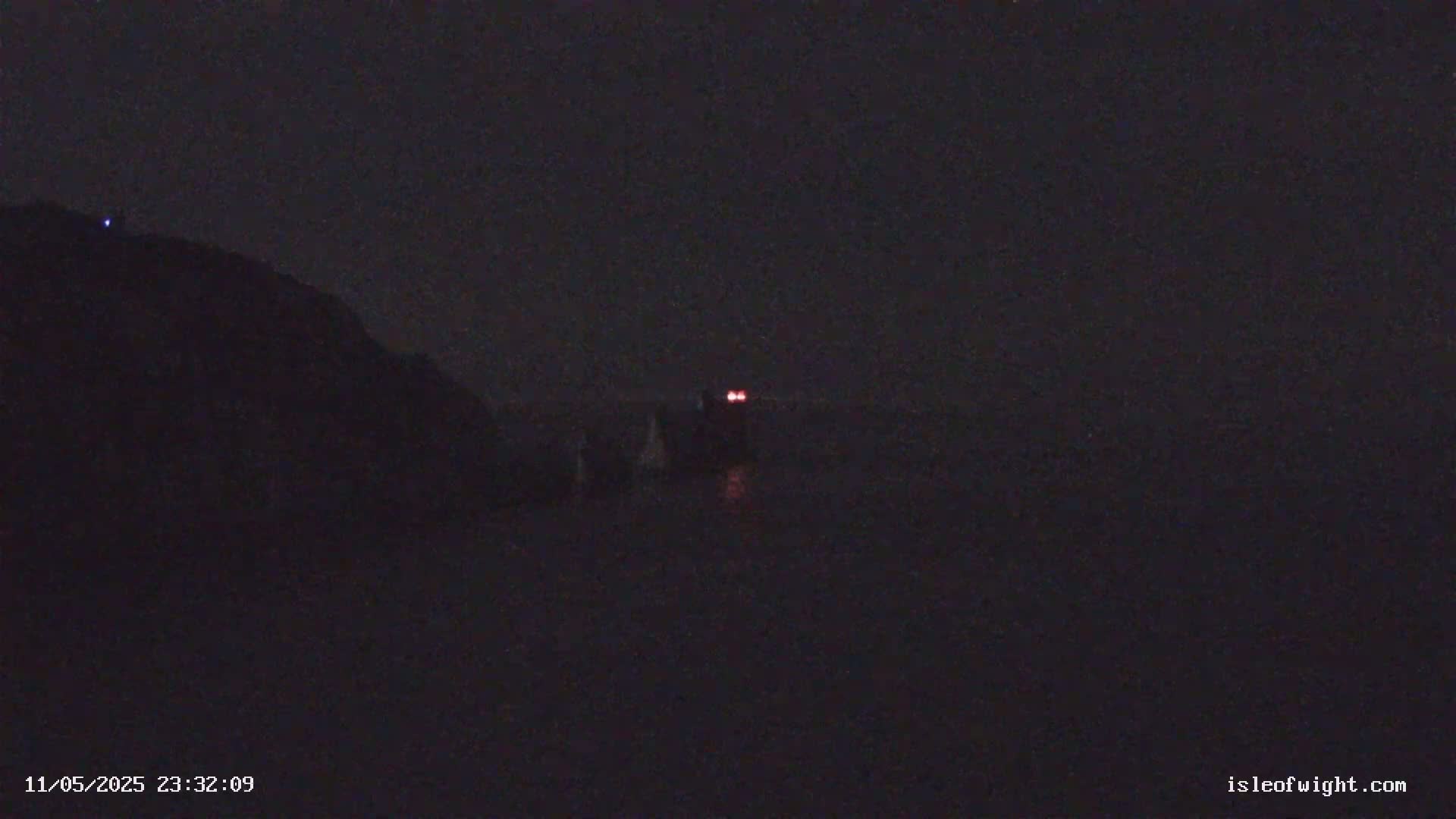 A dark, nighttime scene shows a distant structure with two red lights and a dark hill or cliff in the foreground under a clear, starless sky.