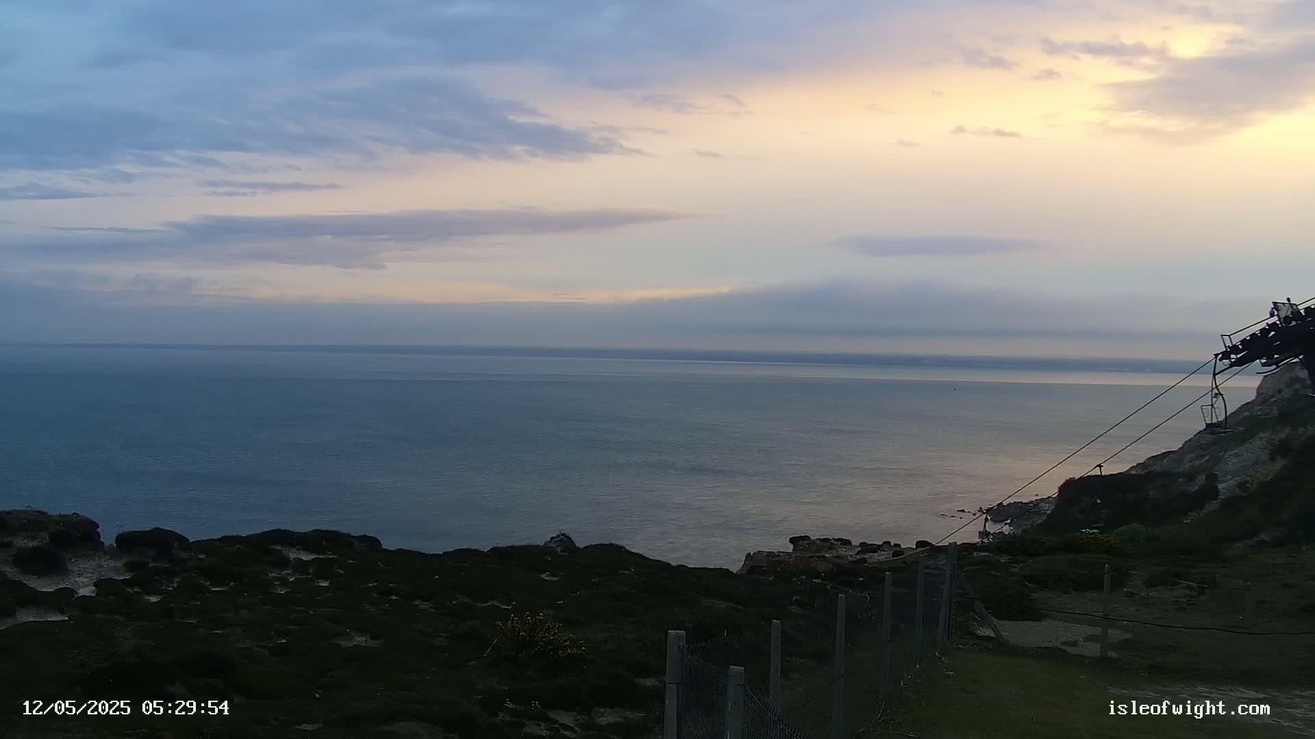 A calm sea stretches to a pale, cloudy sky at sunset, viewed from a clifftop with a chairlift partially visible.