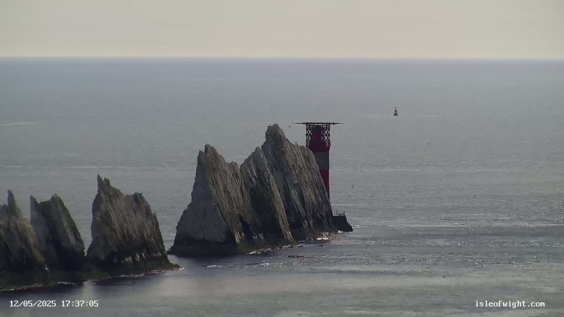 A red and white lighthouse stands atop a cluster of jagged, grey rock formations extending into a calm, grey sea.