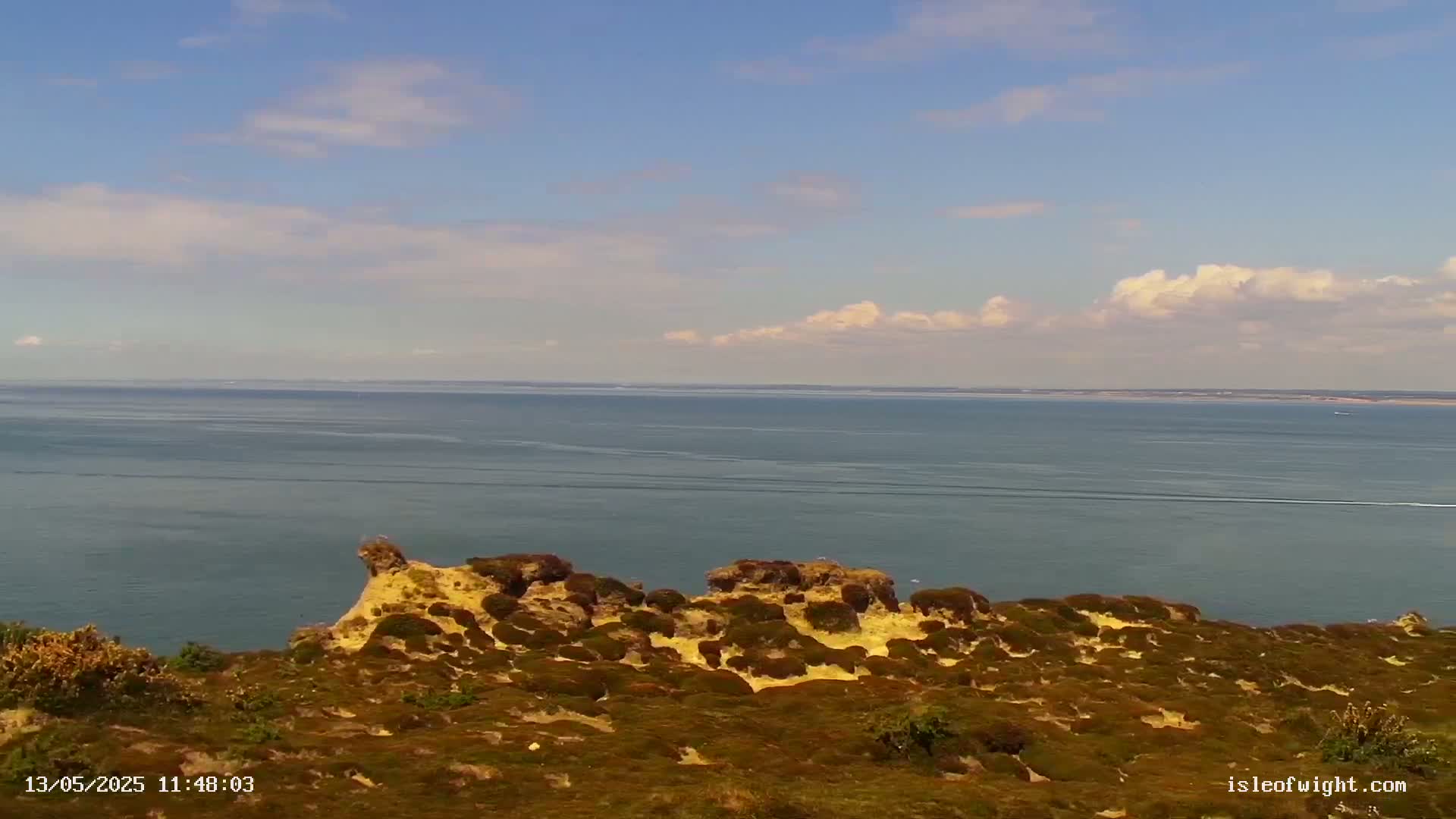 A coastal view shows a calm sea under a partly cloudy sky, with low-lying, scrub-covered, sandy cliffs in the foreground.