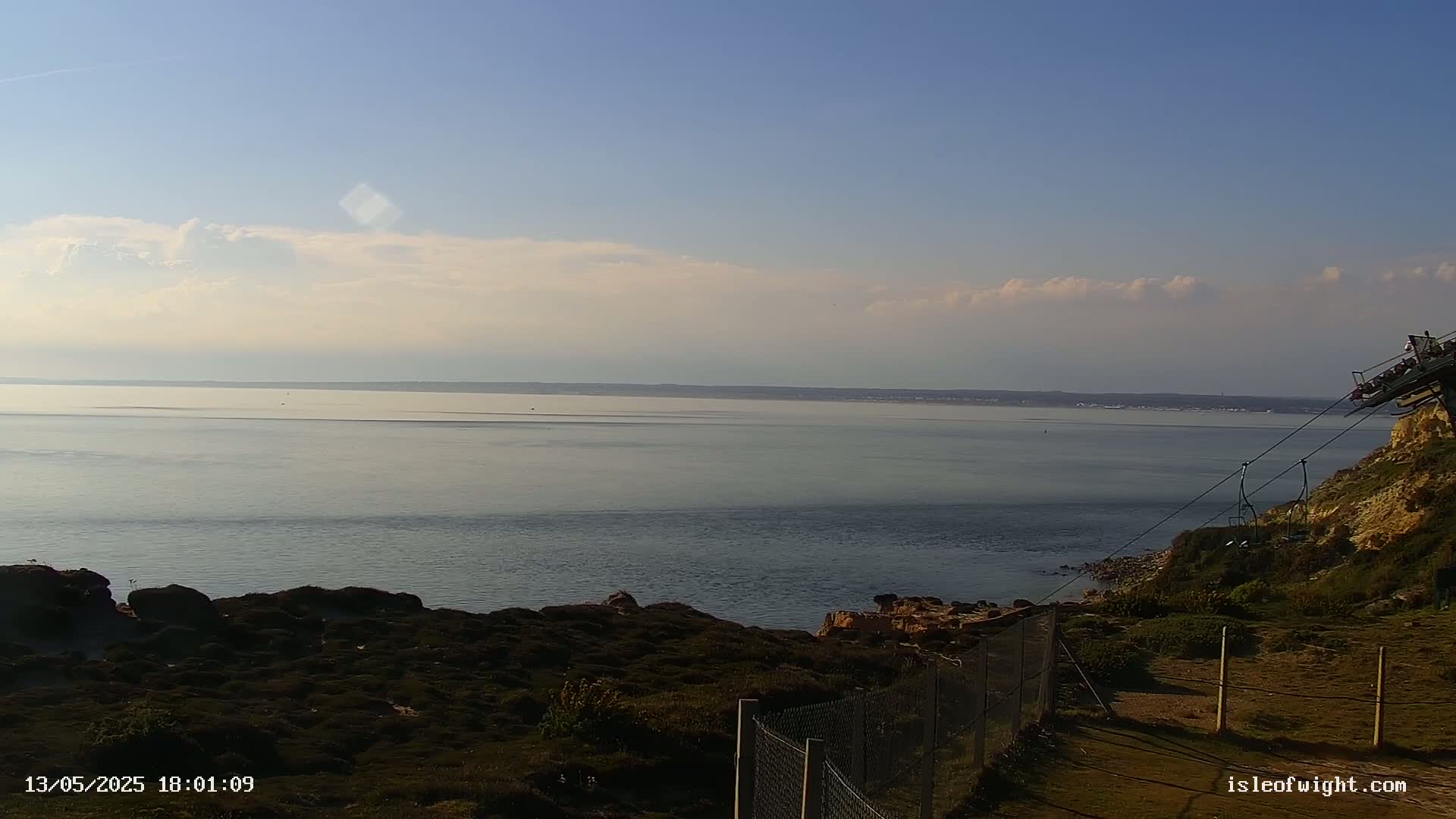 A calm sea stretches to a distant land under a mostly clear sky with a few clouds, viewed from a coastal clifftop with a chairlift partially visible.