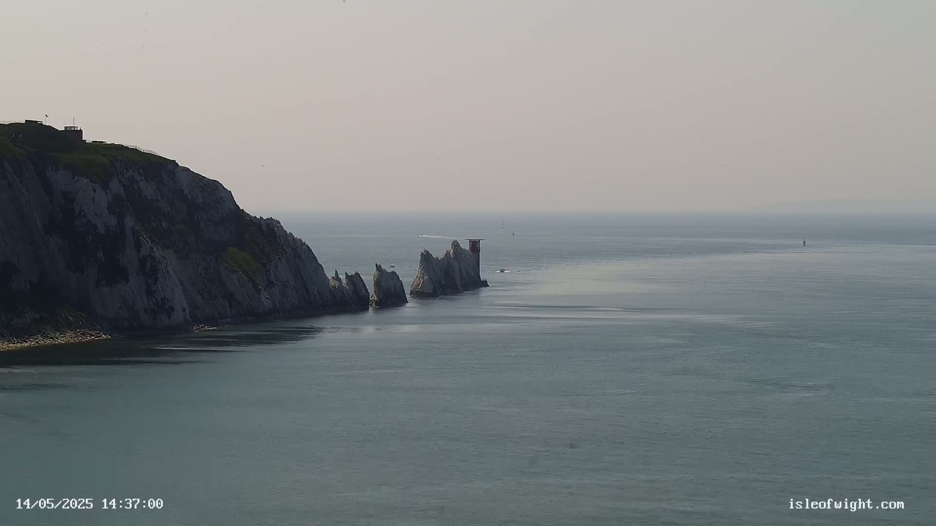 A hazy, coastal scene shows a tall, rocky cliff beside a series of smaller, jagged rocks and a small lighthouse extending into a calm sea.
