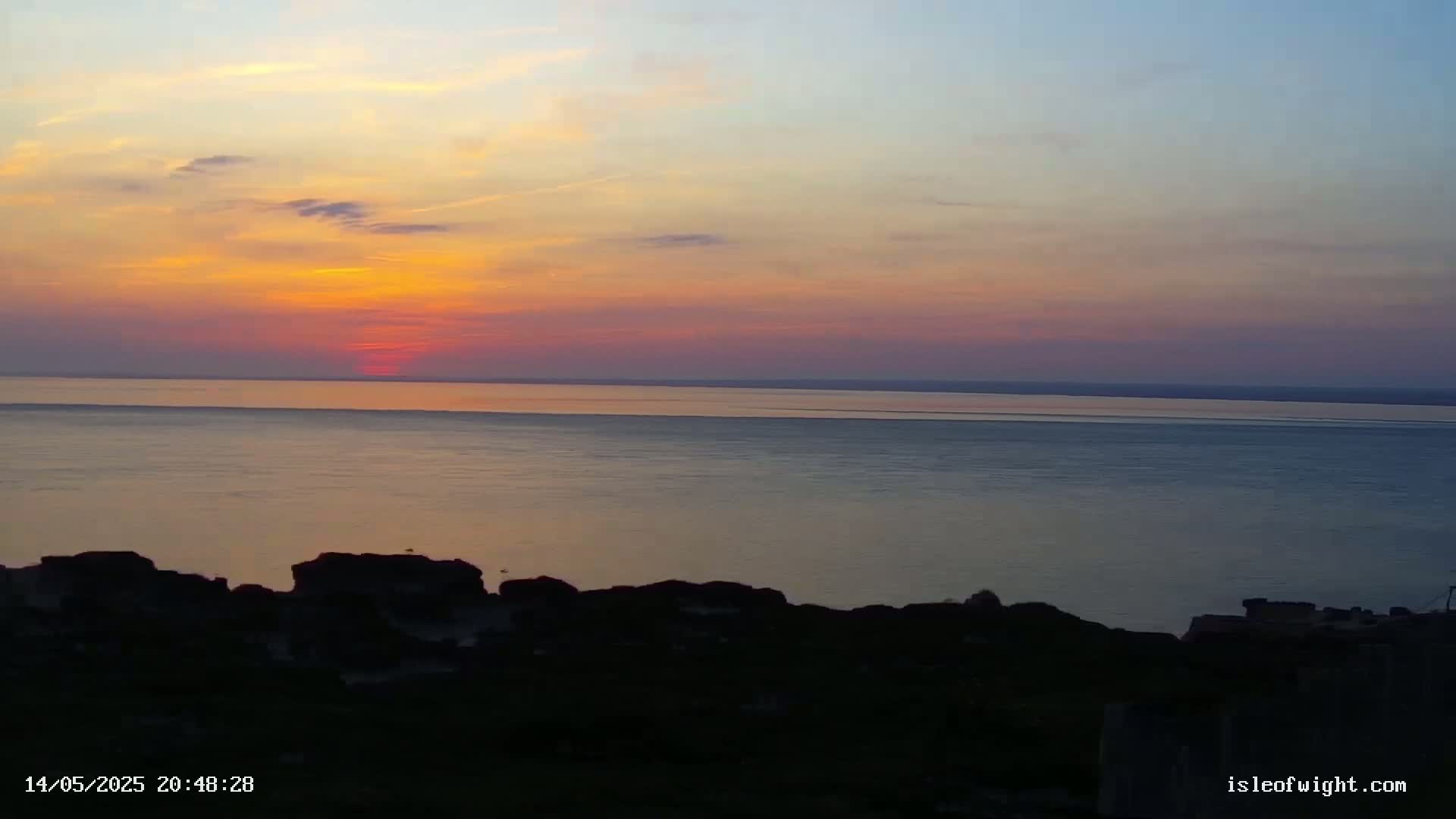 A sunset over a calm body of water, viewed from a rocky, silhouetted shoreline.