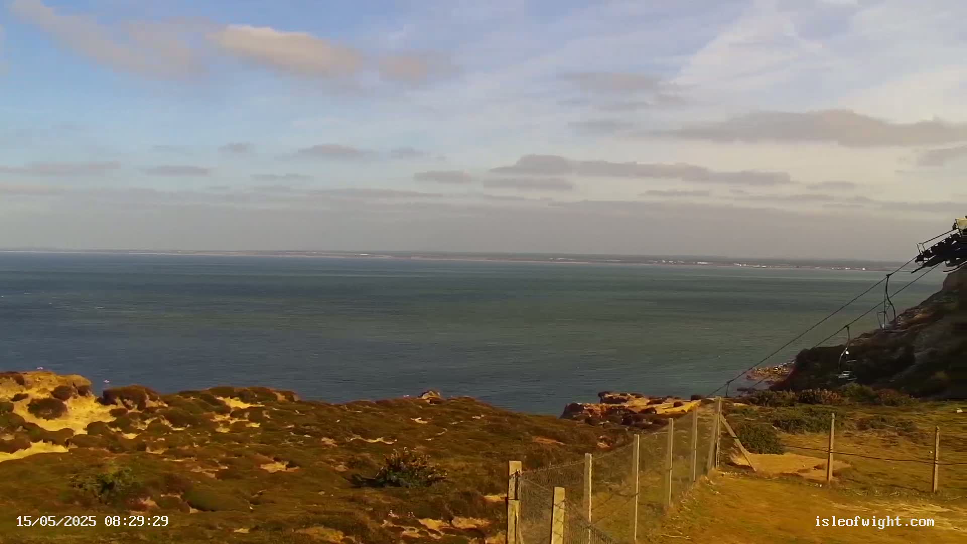 A partly cloudy day reveals a view of a calm sea from a grassy clifftop, fenced and overlooking a vast expanse of water.