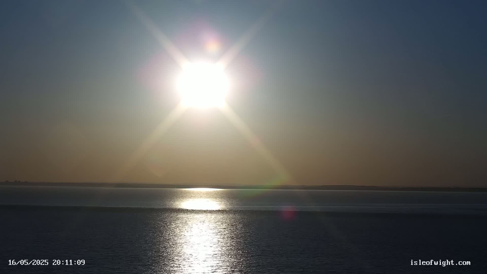 A bright sun shines over a calm body of water with a distant shoreline.