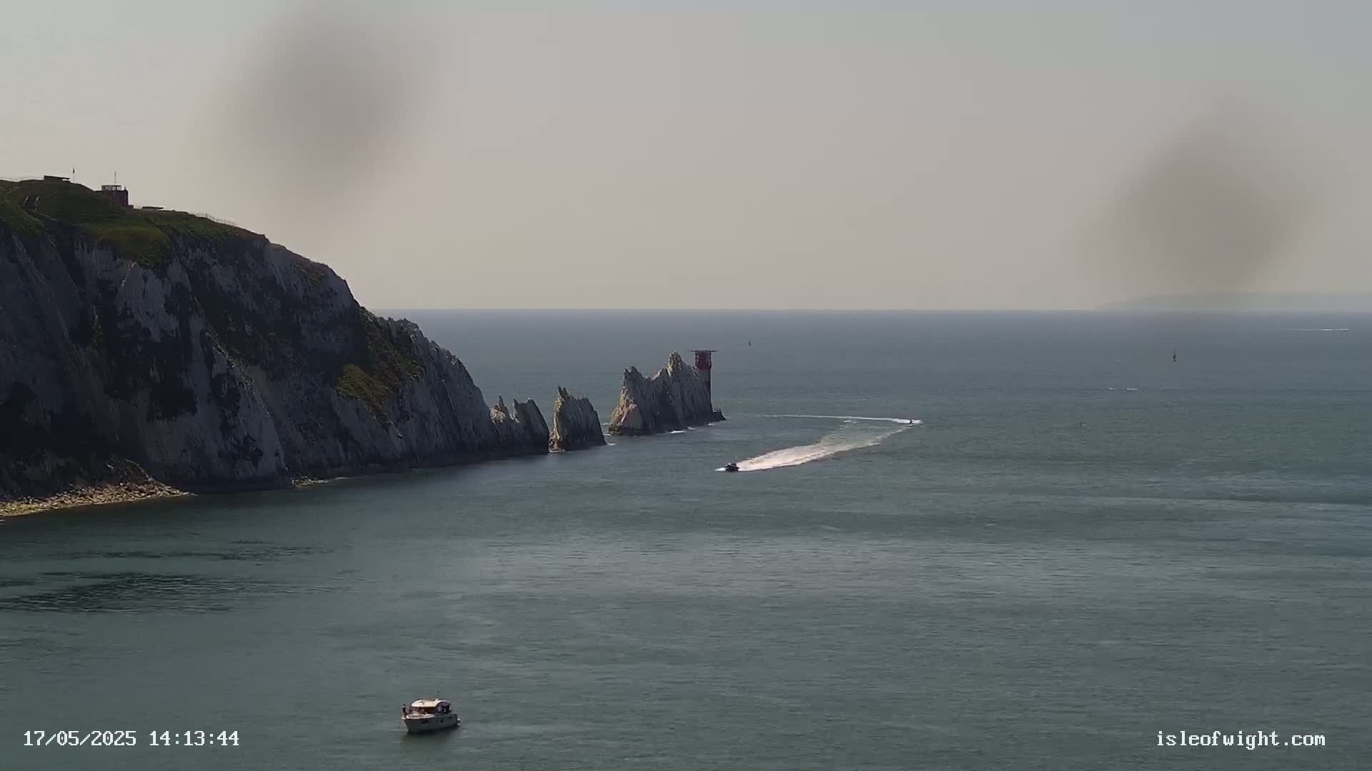 A hazy day reveals a rocky coastline with a lighthouse and two small boats on calm waters.
