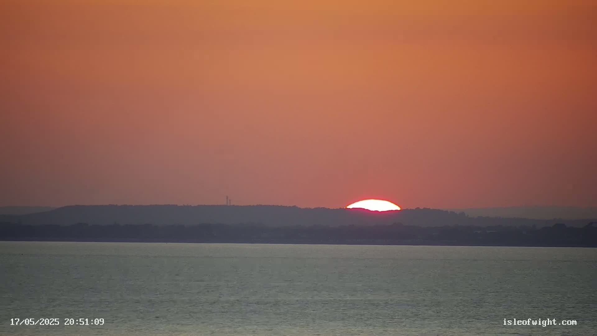 A large, bright sun sets behind a dark landmass over a calm body of water under a reddish-orange sky.