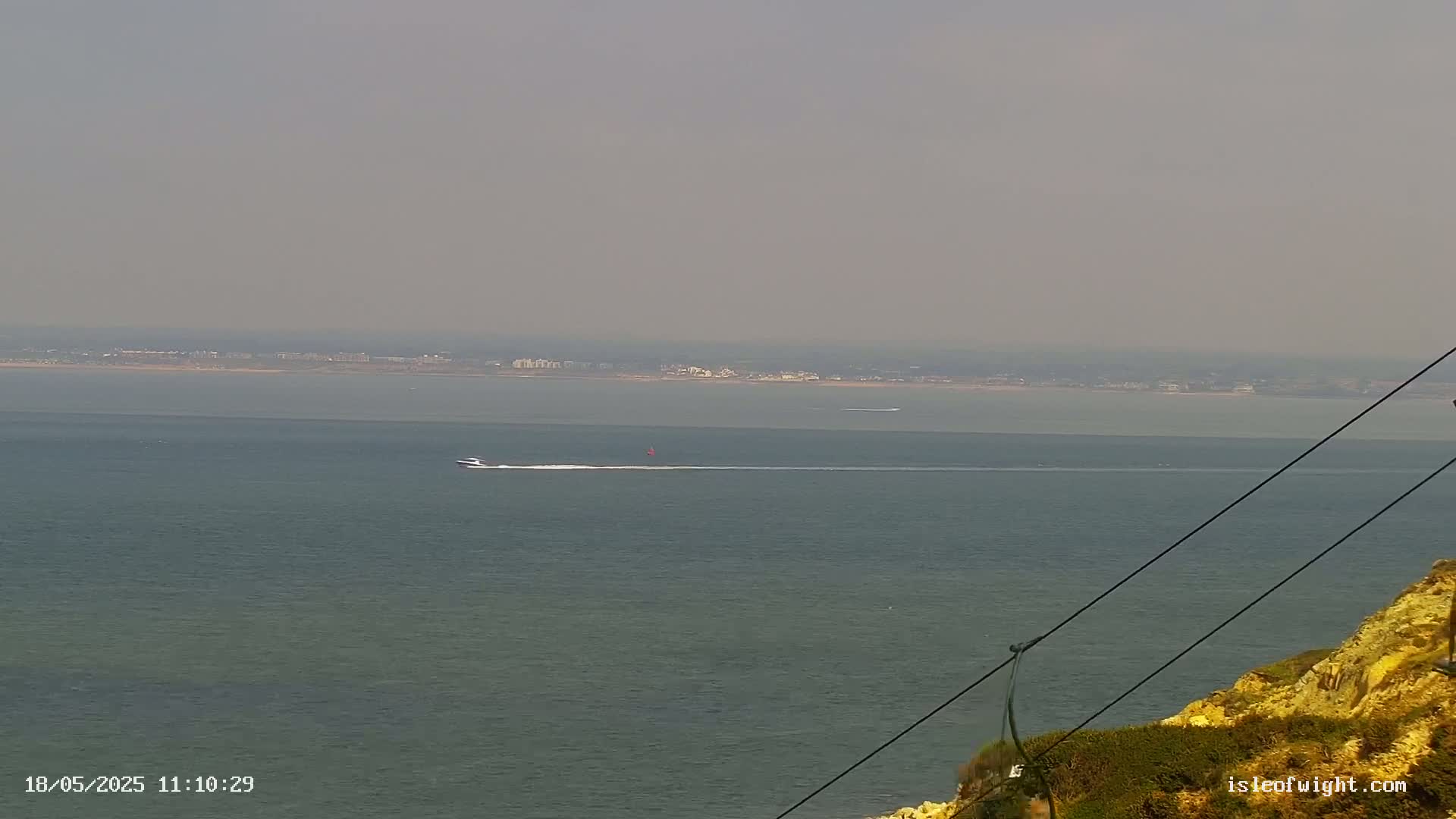 A hazy view of a calm sea with a small boat and distant shoreline is seen from a high vantage point, with cables in the foreground.