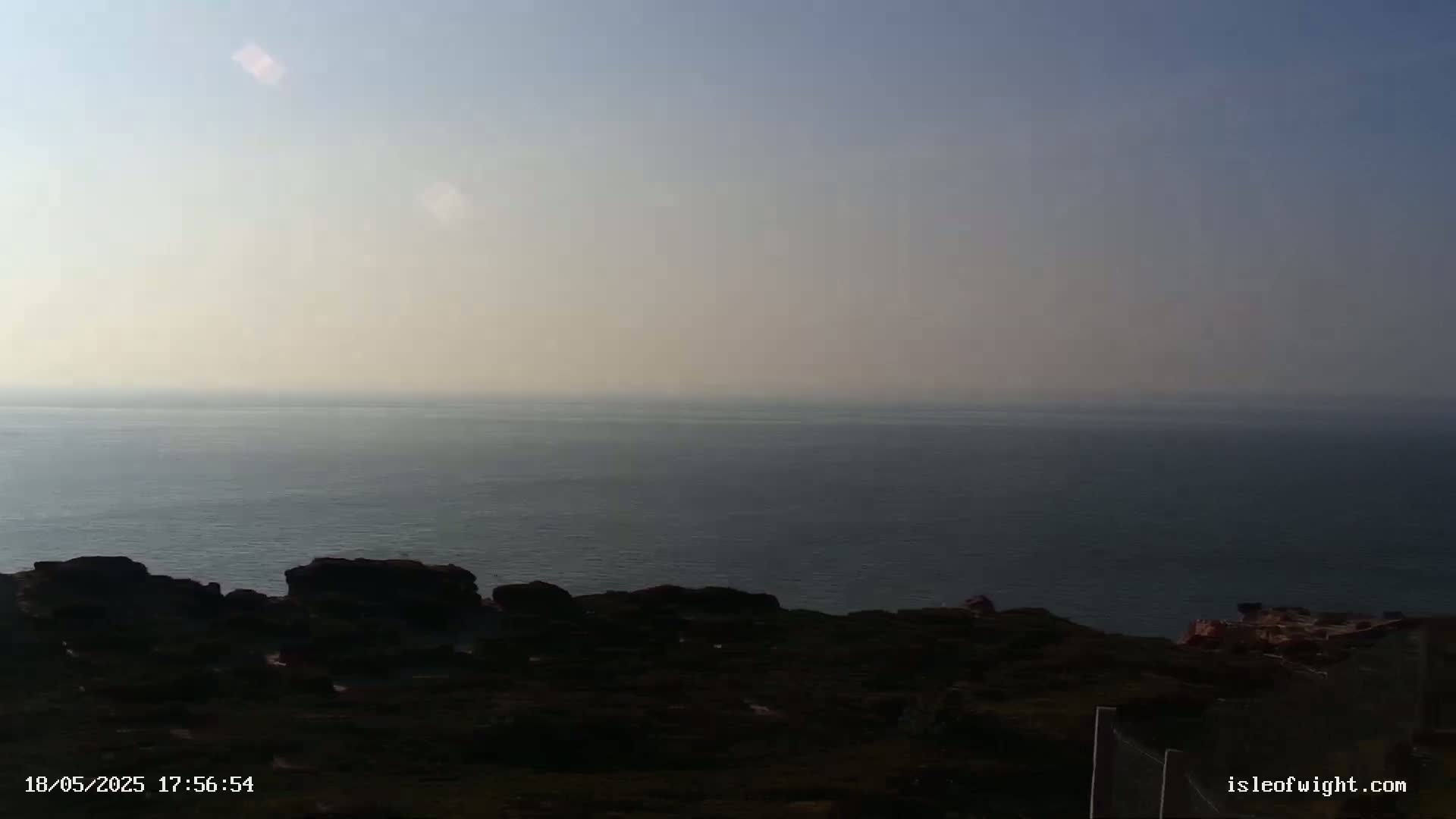 A hazy, calm ocean stretches to the horizon from a dark, rocky coastline under a clear, light-blue sky.