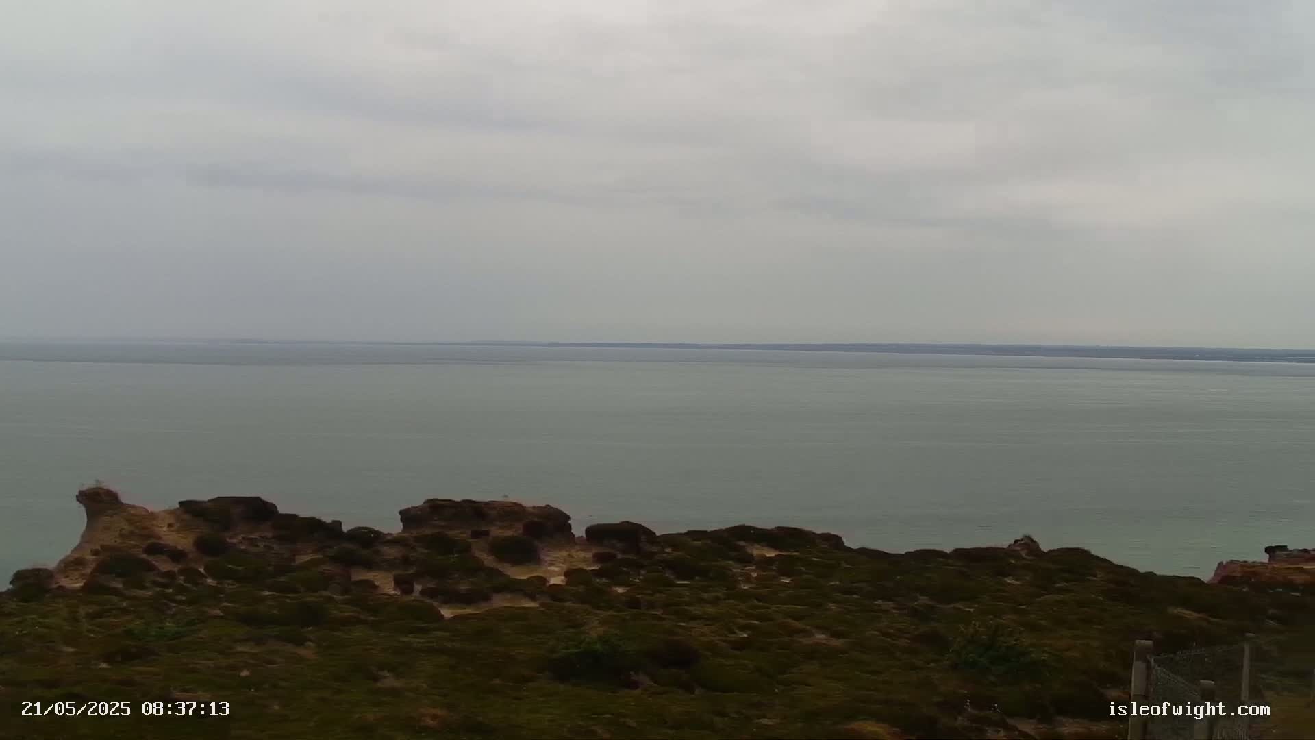 A rocky, shrub-covered cliff overlooks a calm, gray sea under an overcast sky.