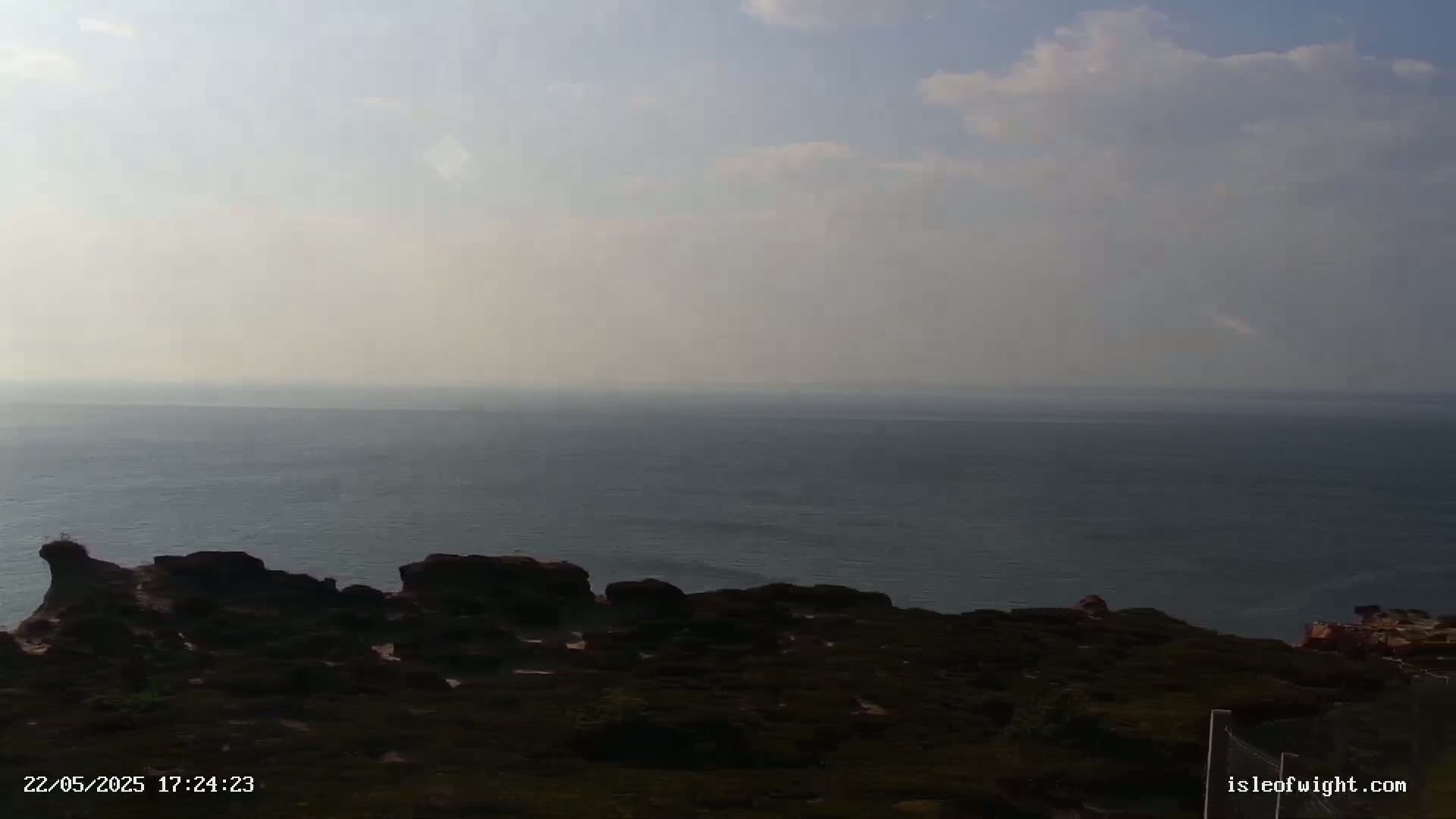 A hazy, partly cloudy sky overlooks a dark-colored, rocky shoreline and a calm, grey-blue sea.