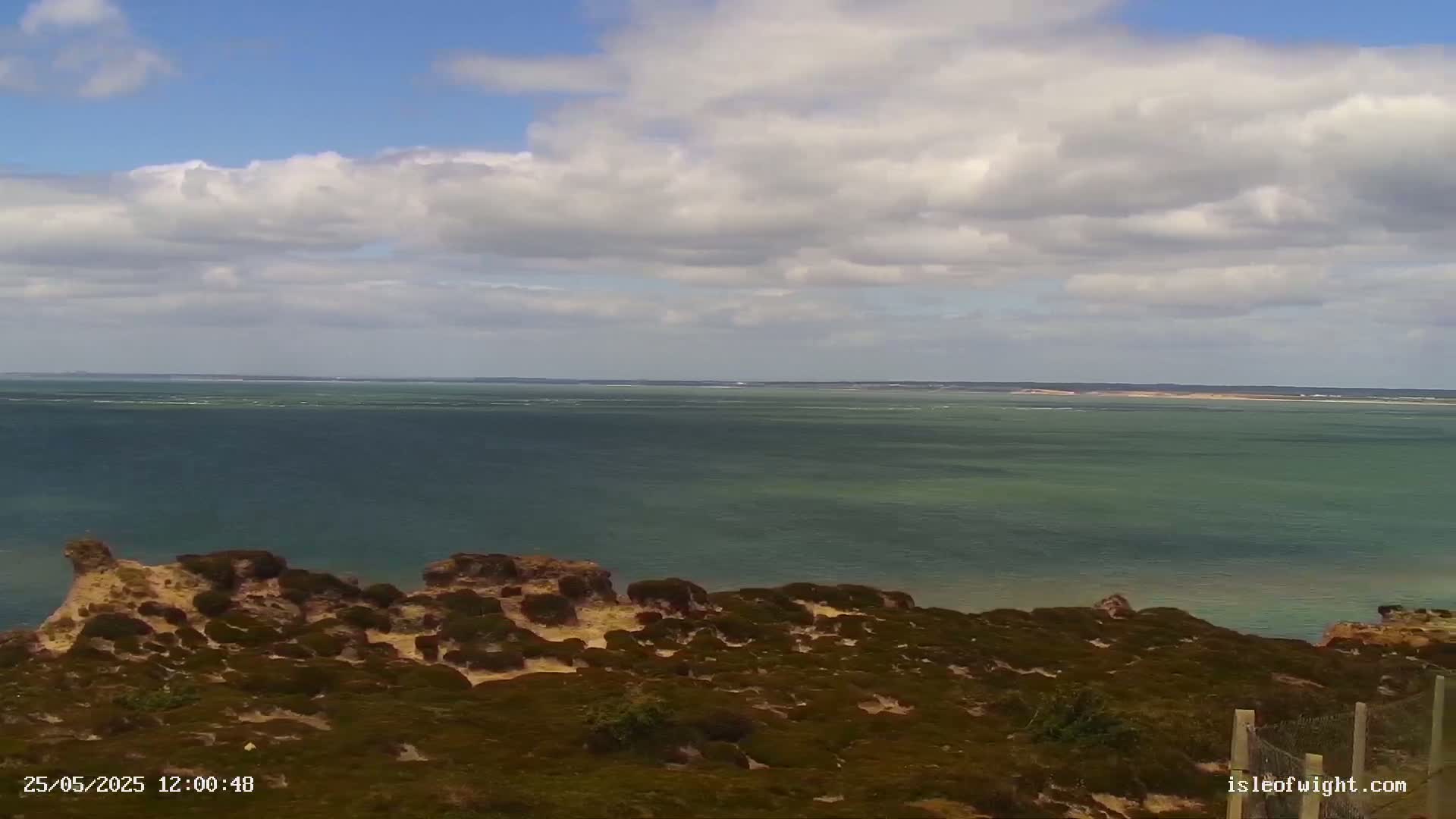 A partly cloudy day shows a view of a calm, greenish-blue sea from a coastal clifftop covered in low-lying, brown vegetation.