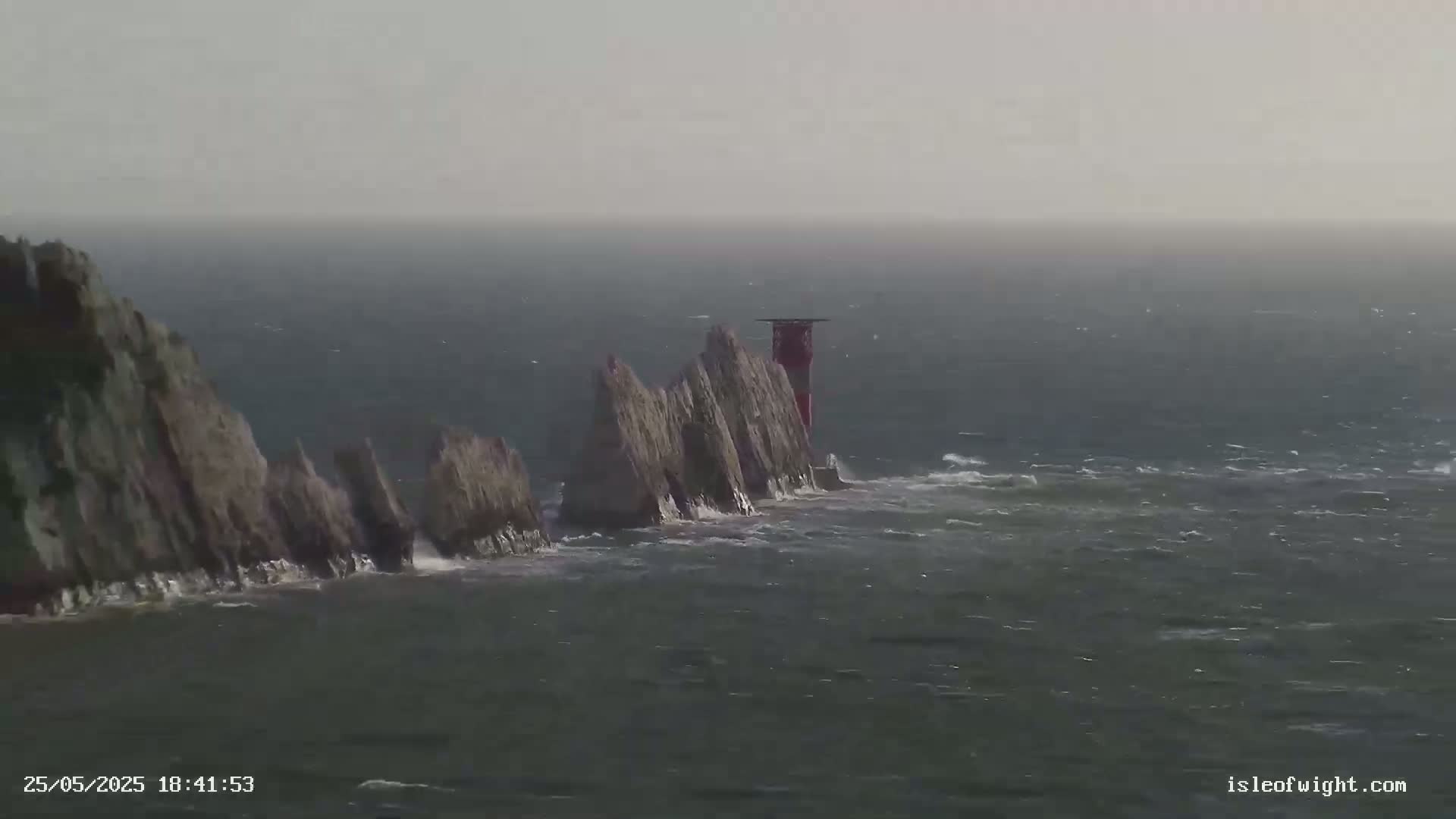 A series of white rock formations with a red and white lighthouse at the end are battered by waves in hazy weather conditions.