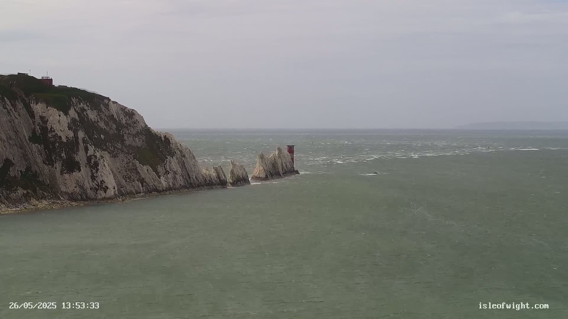 A white cliff face juts into a choppy, grey sea where a line of smaller white rock formations extends from it towards a small red and white lighthouse.