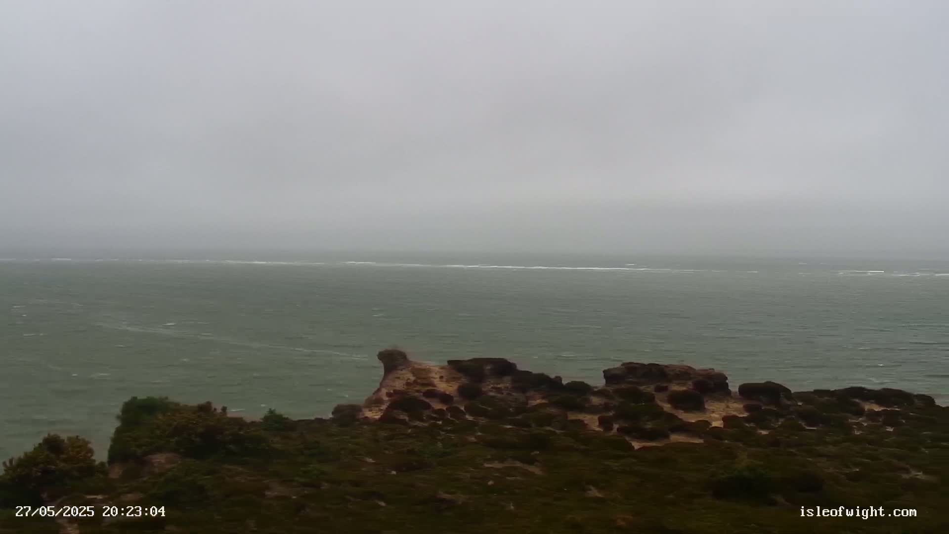 A view from a clifftop shows a grey, overcast sea with small waves under a foggy sky.