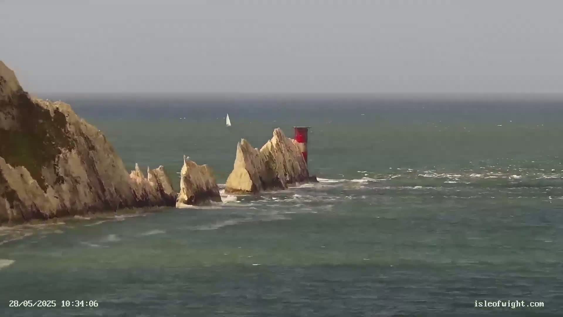 A small sailboat is visible on a somewhat choppy sea, near a rocky outcrop with a red and white lighthouse, and a larger cliff face.
