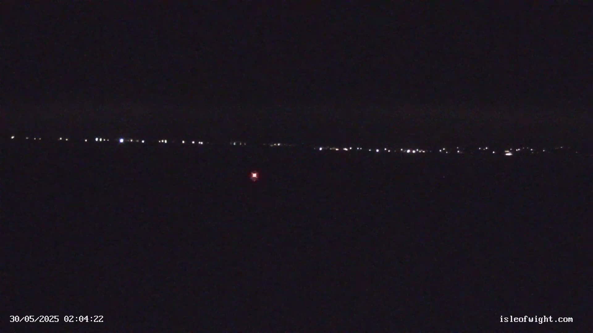 A dark, nighttime scene shows a distant line of small, faint lights across a black horizon, with a single, brighter red light closer to the bottom.
