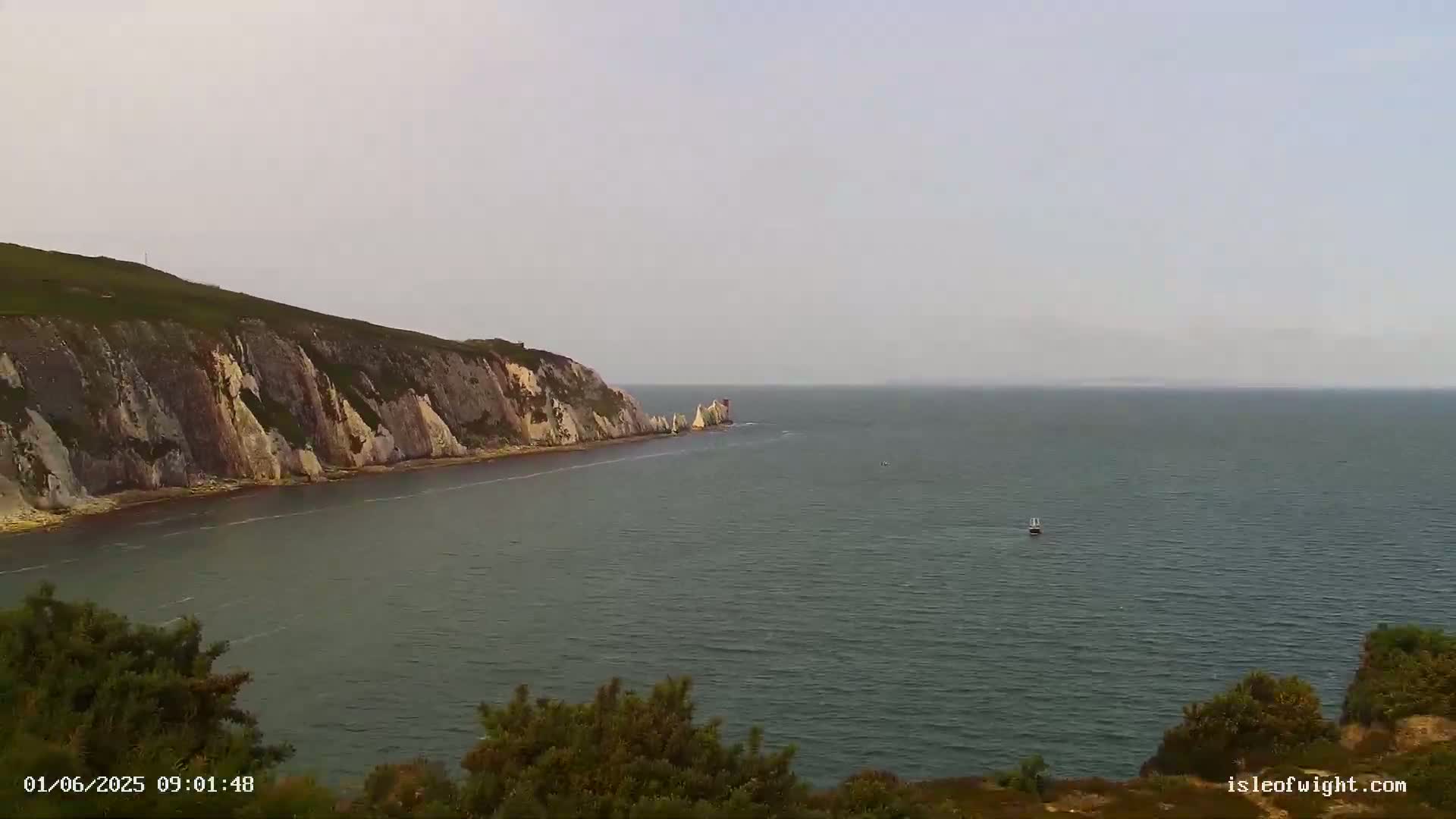 A hazy view of a calm sea shows a coastal cliff face with layered rock formations extending into the water, with a small boat visible in the distance.
