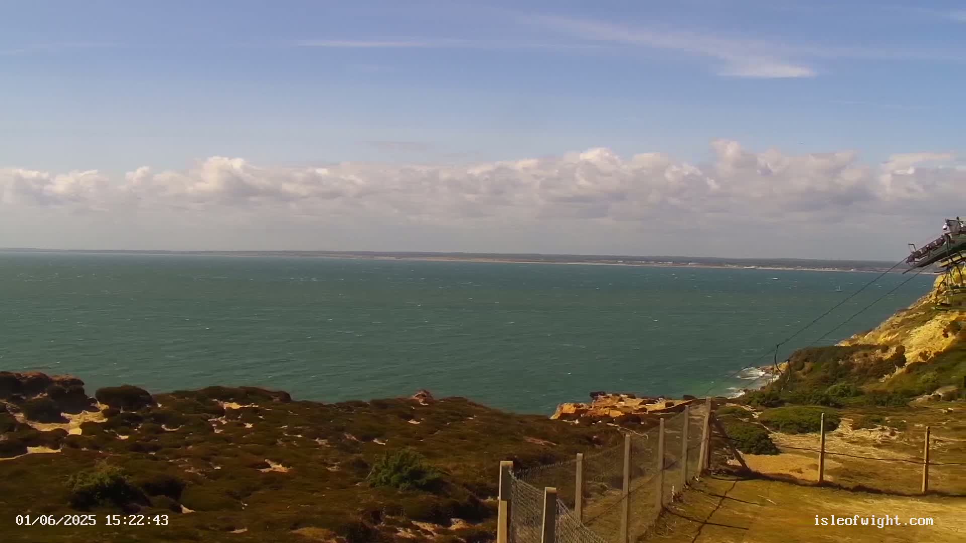 A partly cloudy day reveals a view of a calm sea from a clifftop, with low-lying vegetation and a wire fence in the foreground.