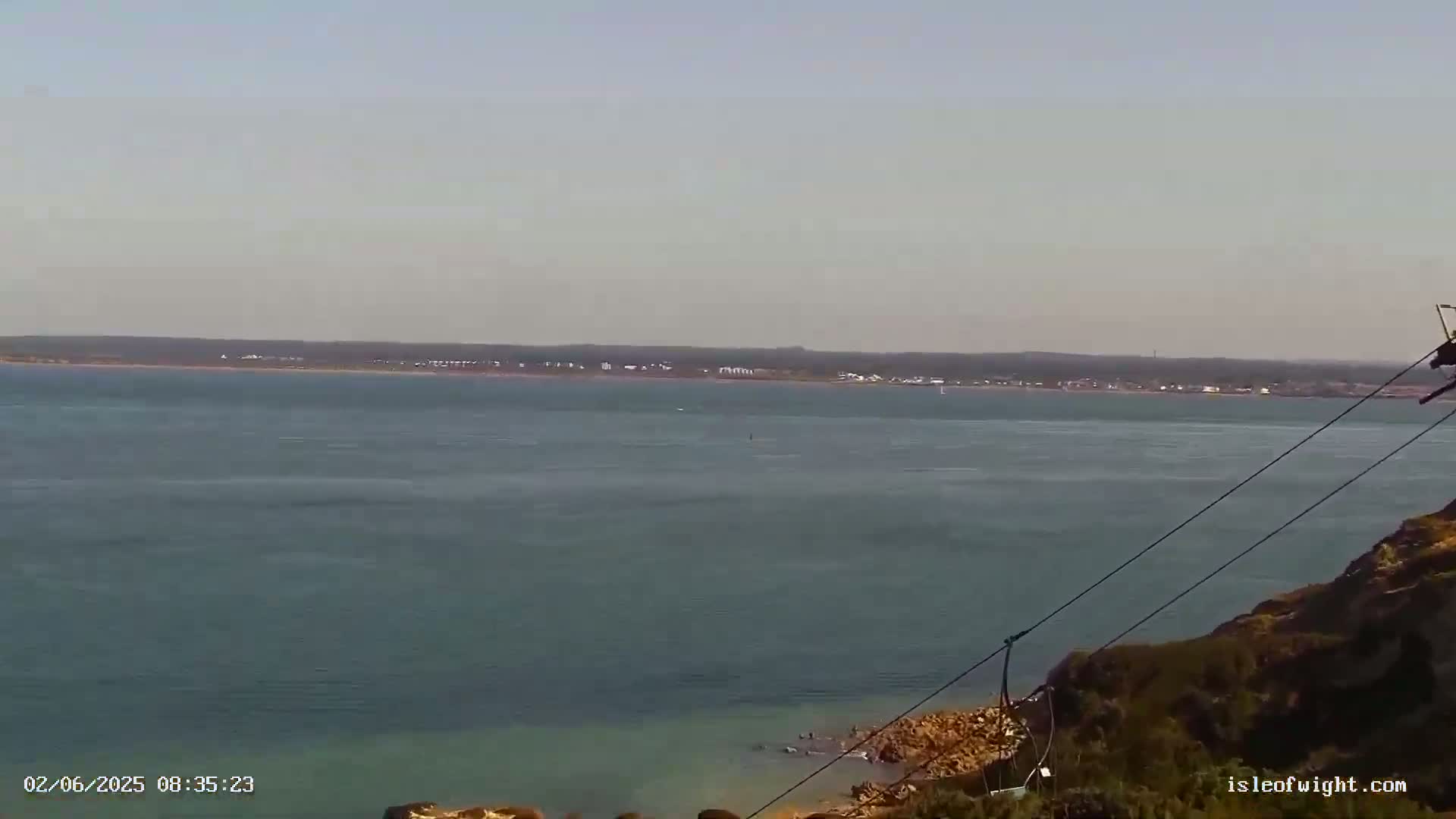 A calm body of water stretches to a distant shoreline under a clear sky, with a portion of a hillside and what appears to be a zip line in the foreground.