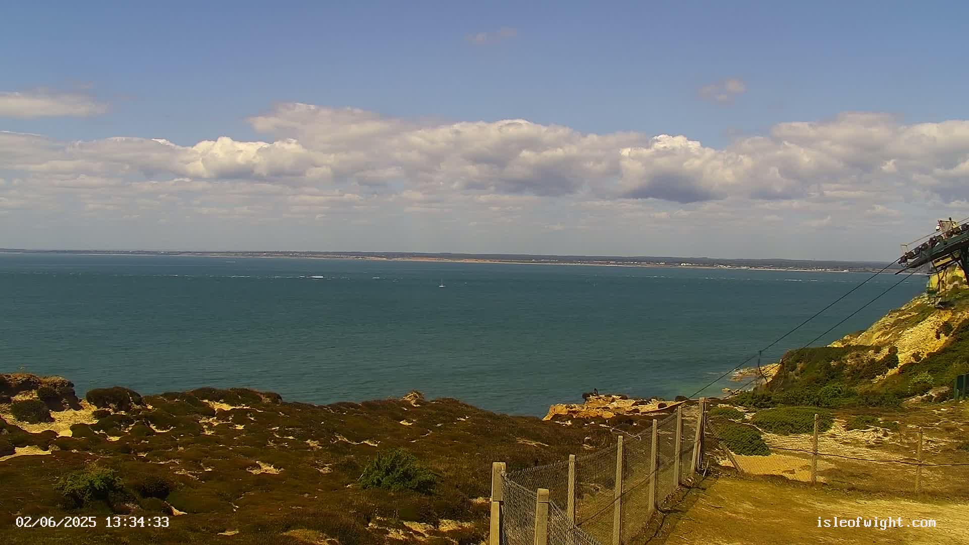A partly cloudy day shows a view of a calm sea from a clifftop, with scrubby vegetation and a wire fence in the foreground.
