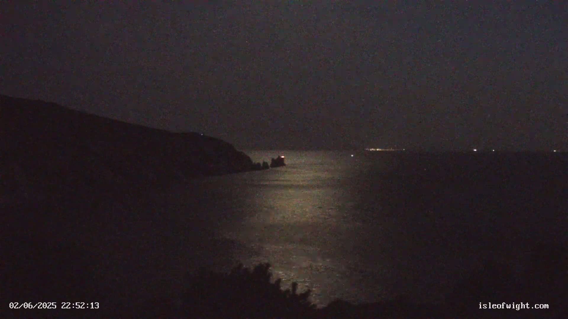 A dark, moonlit night scene shows a body of water reflecting the moon's light, with a distant shoreline and a small, lit structure on a rocky outcrop in the water.