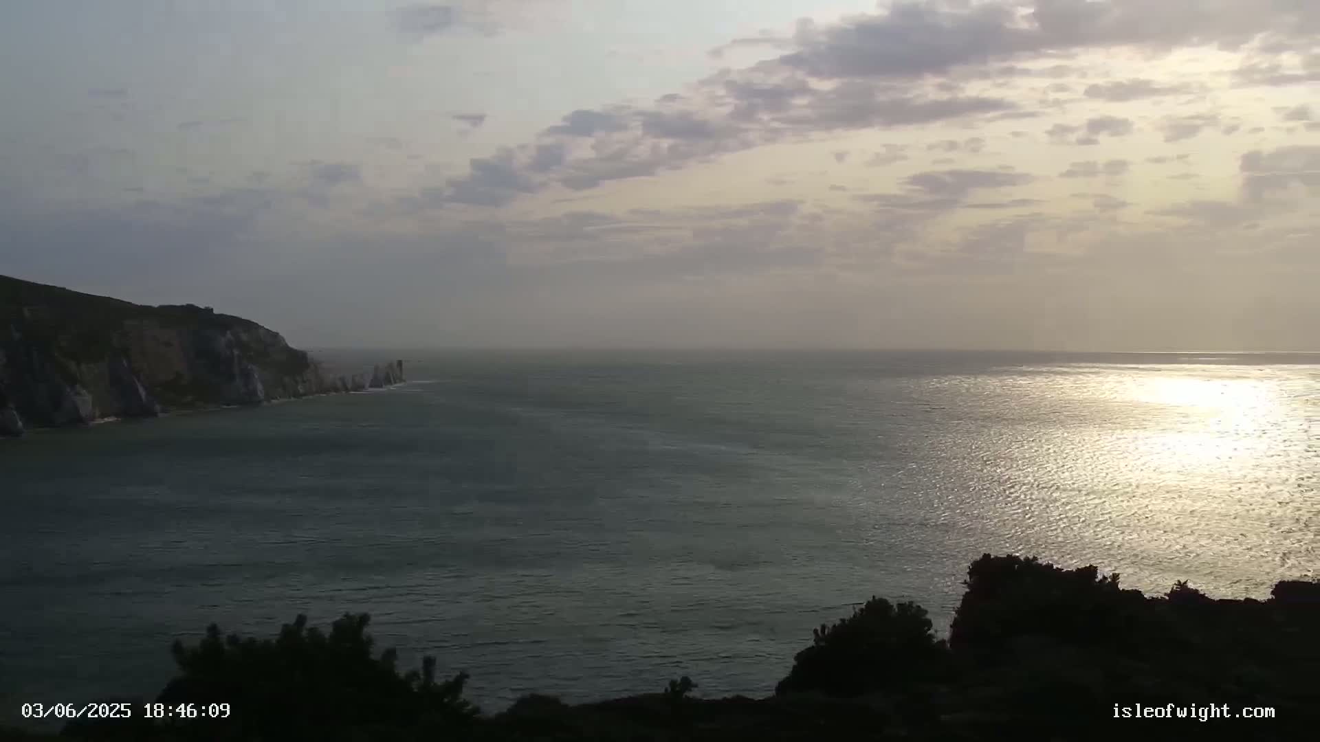 A calm sea reflects the setting sun under a partly cloudy sky, with a cliff face visible in the distance.