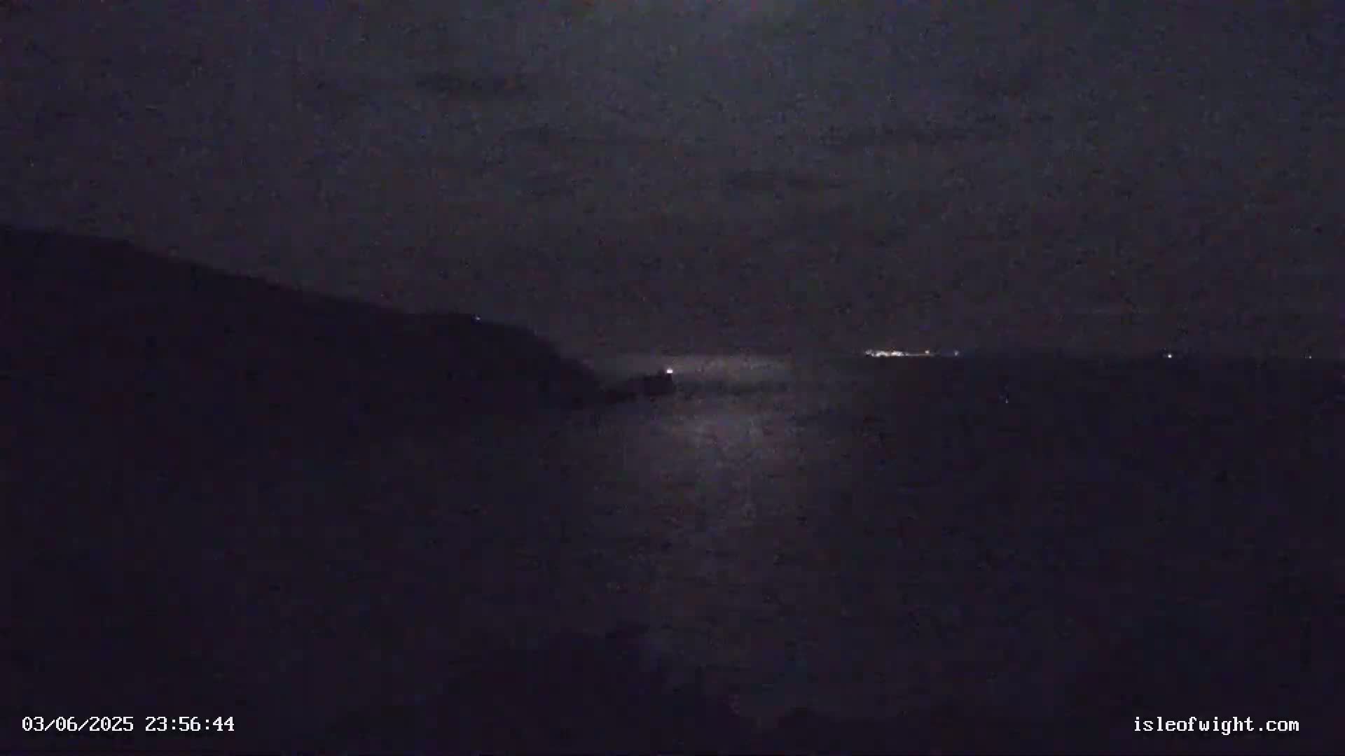 A dark, nighttime scene shows a body of water reflecting a bright light source, with a dark landmass to the left and faint lights visible in the distance across the water.