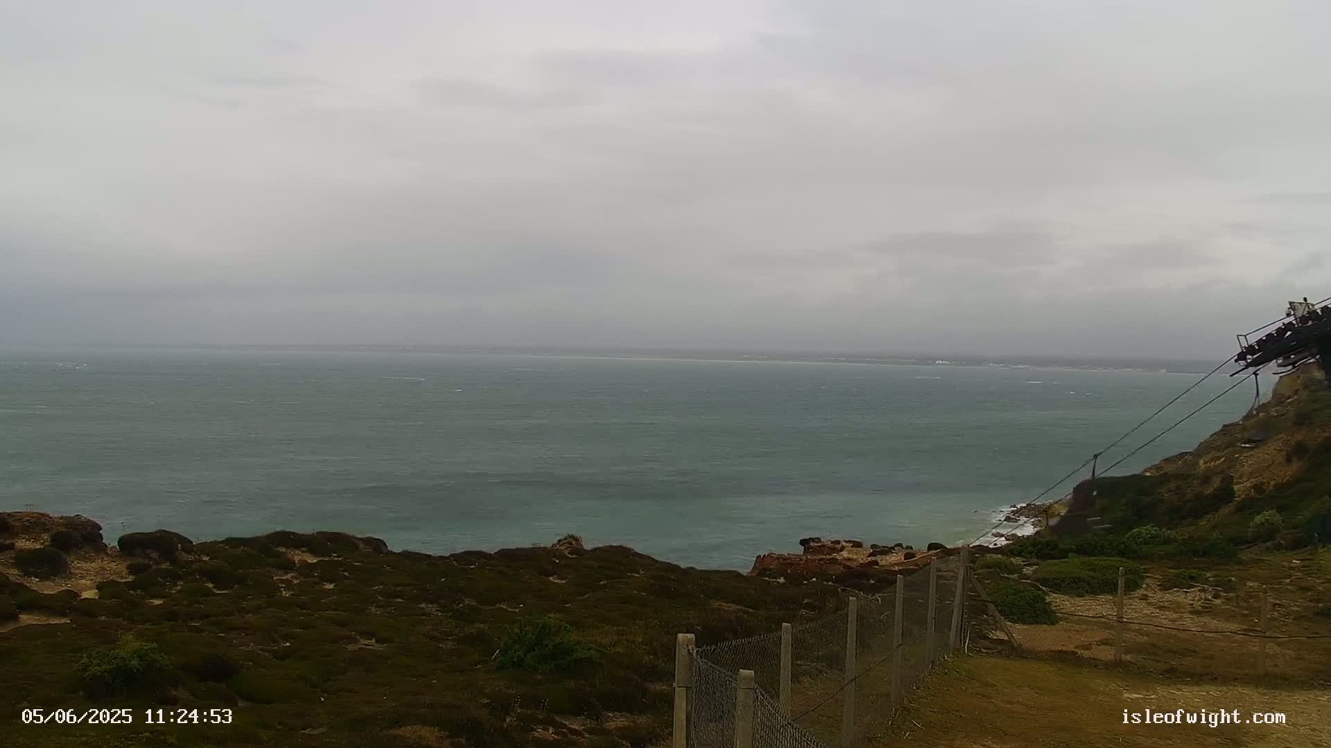 A grey, overcast day shows a dark-green ocean stretching to a hazy horizon, viewed from a rocky, shrub-covered clifftop with a chain-link fence in the foreground.