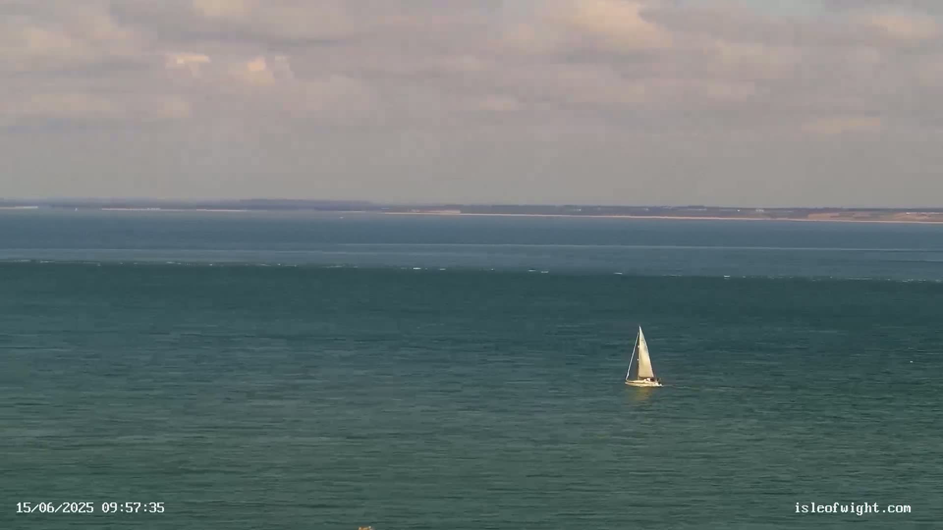A sailboat is visible on a calm, dark-green sea under a mostly cloudy sky with a distant landmass on the horizon.
