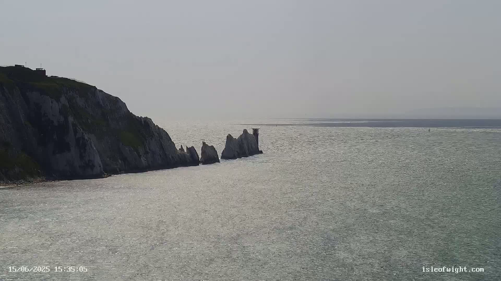A bright, hazy day reveals a rocky cliffside meeting a calm sea, where a line of jagged rocks extends into the water.