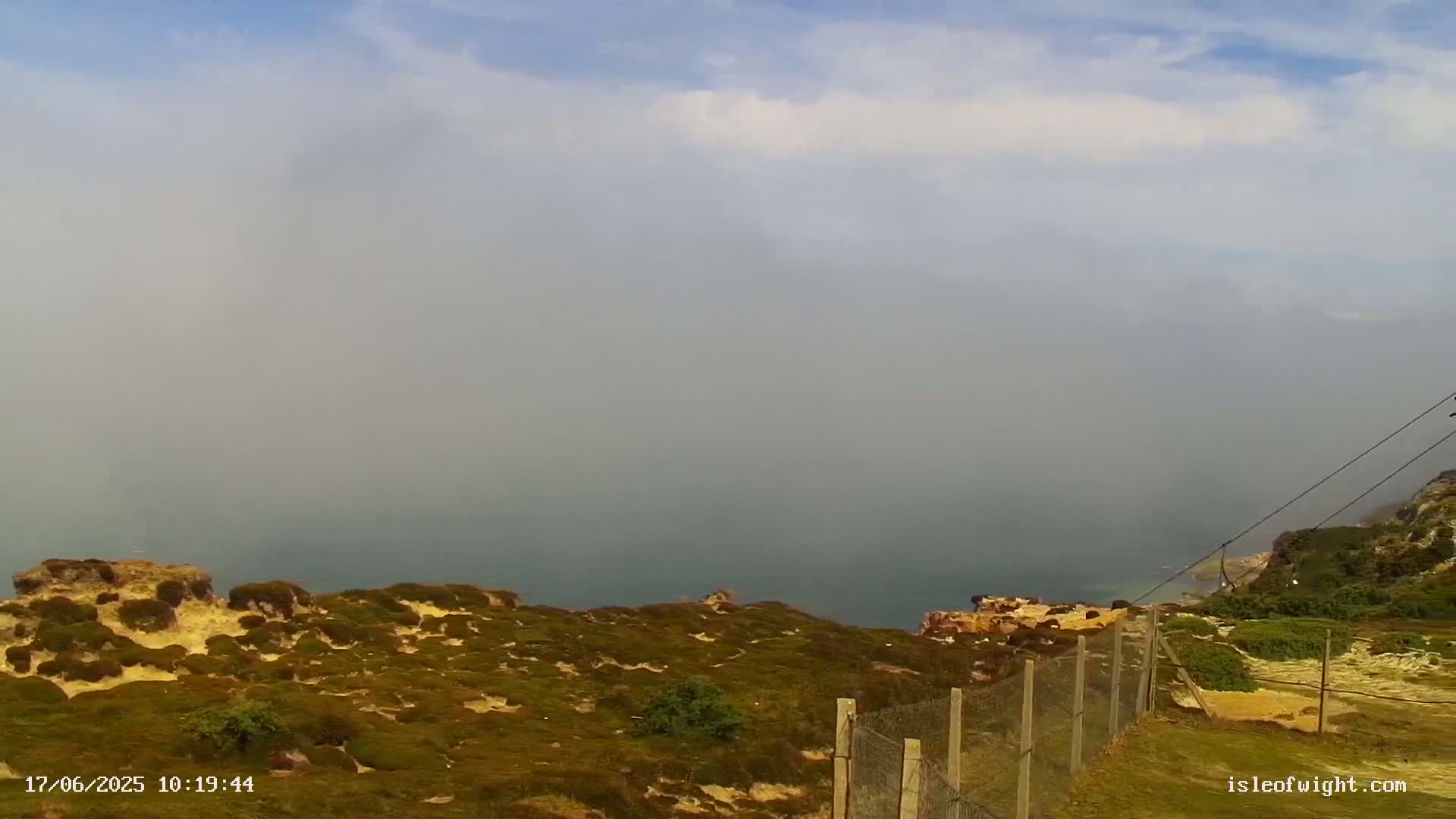 A hazy, foggy day overlooks a coastal area with low-lying vegetation and a wire fence.
