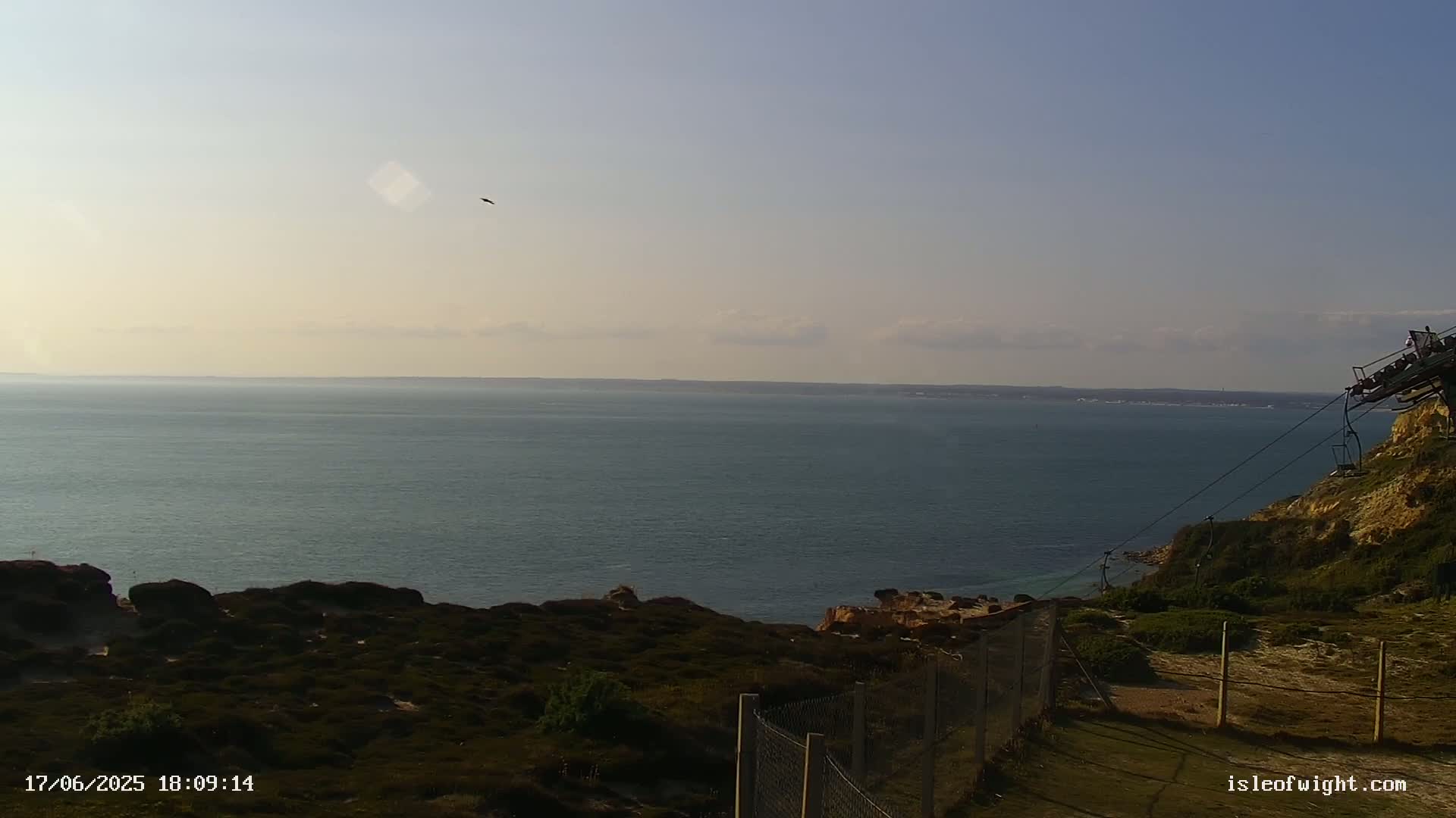 A calm sea stretches to the horizon under a mostly clear sky, viewed from a coastal cliff with sparse vegetation and a chairlift visible in the foreground.